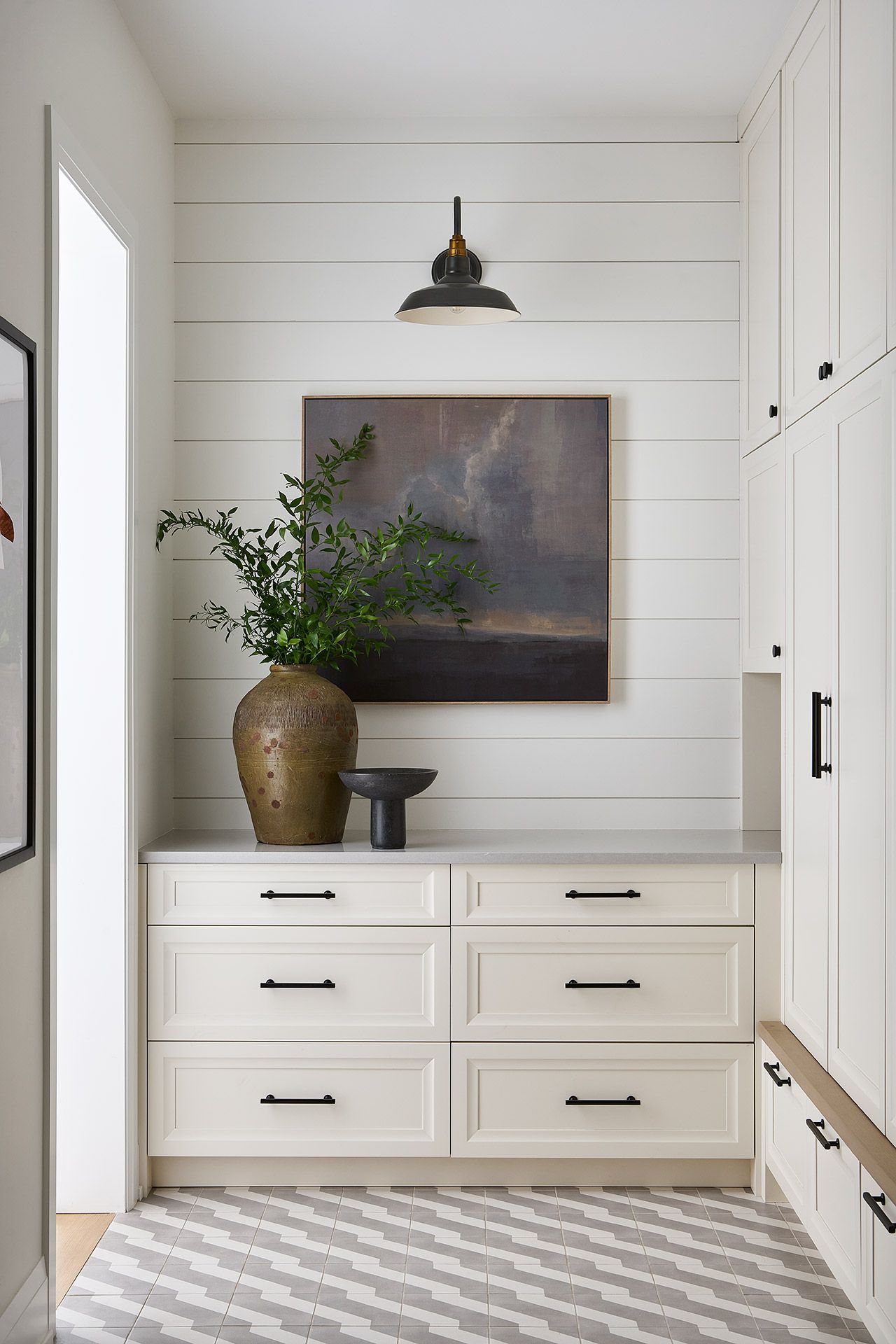Bright hallway with white built-in cabinets, a large vase, potted plant, framed art, and pendant light
