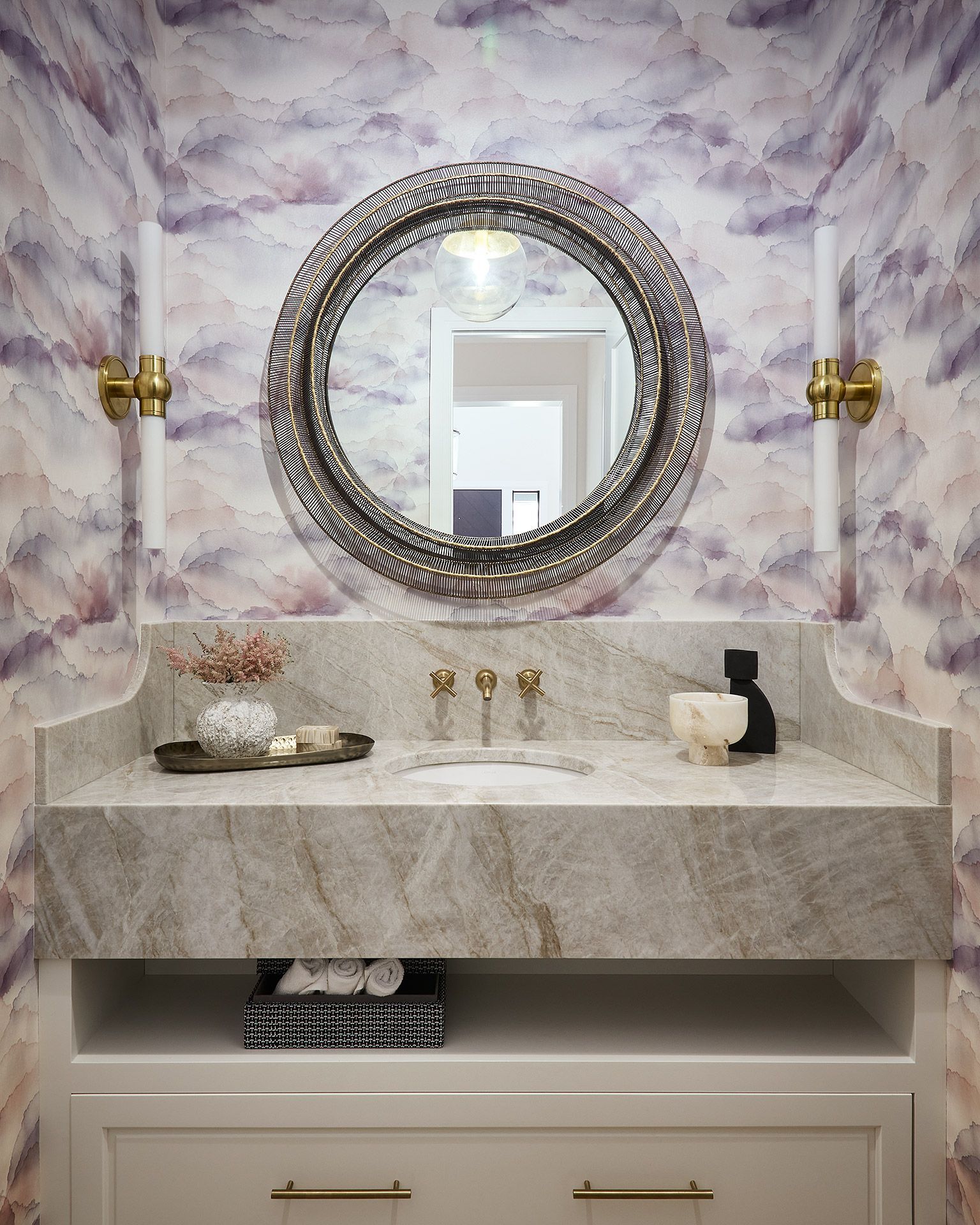Elegant bathroom vanity with marble wall, round ornate mirror, and brass sconces.
