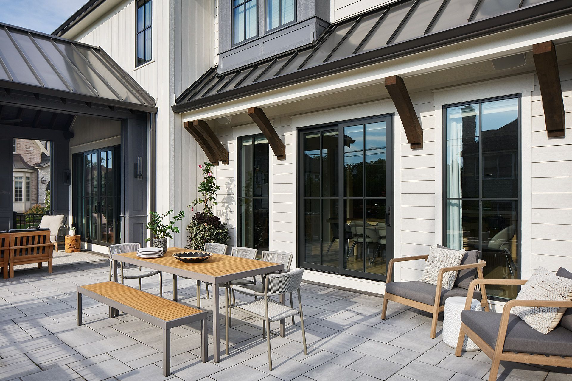 Modern patio with outdoor seating, wood table, and large windows beside a white house