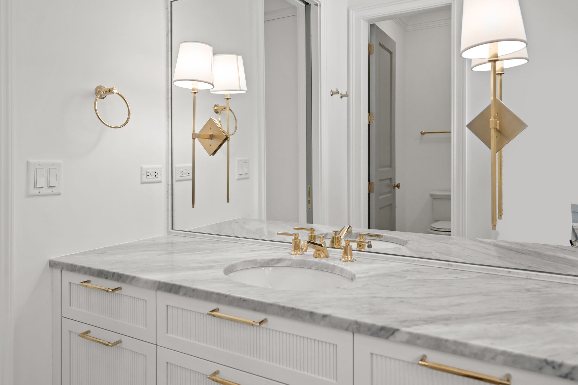 Bright white bathroom vanity with marble countertop, gold faucets, mirror, and wall sconces