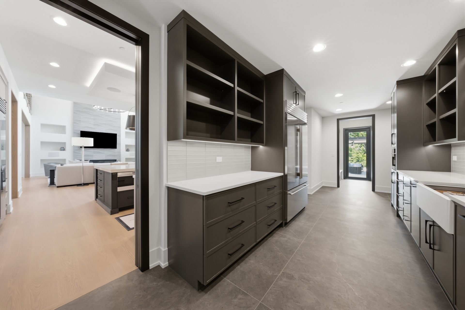 Modern kitchen with dark cabinets, gray tile floor, white countertops, and a view into a living area