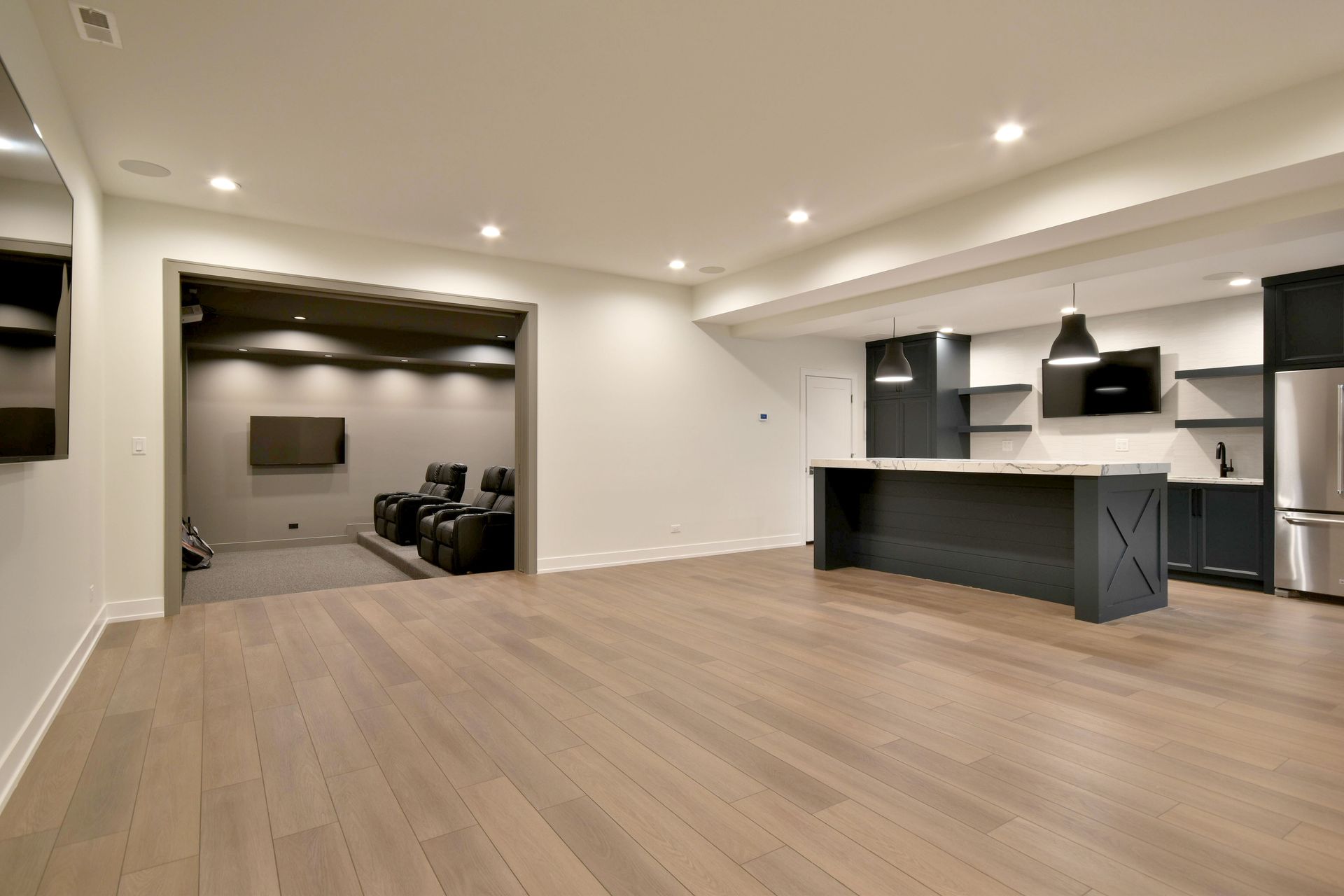 Empty modern basement with wood floors, recessed lights, and a kitchen bar area.