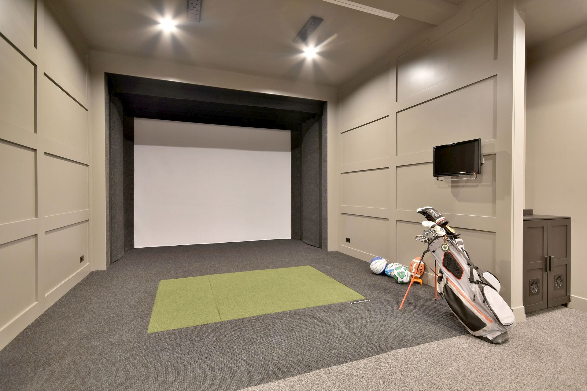 Indoor golf simulator room with a screen, green hitting mat, and golf bags on the floor.