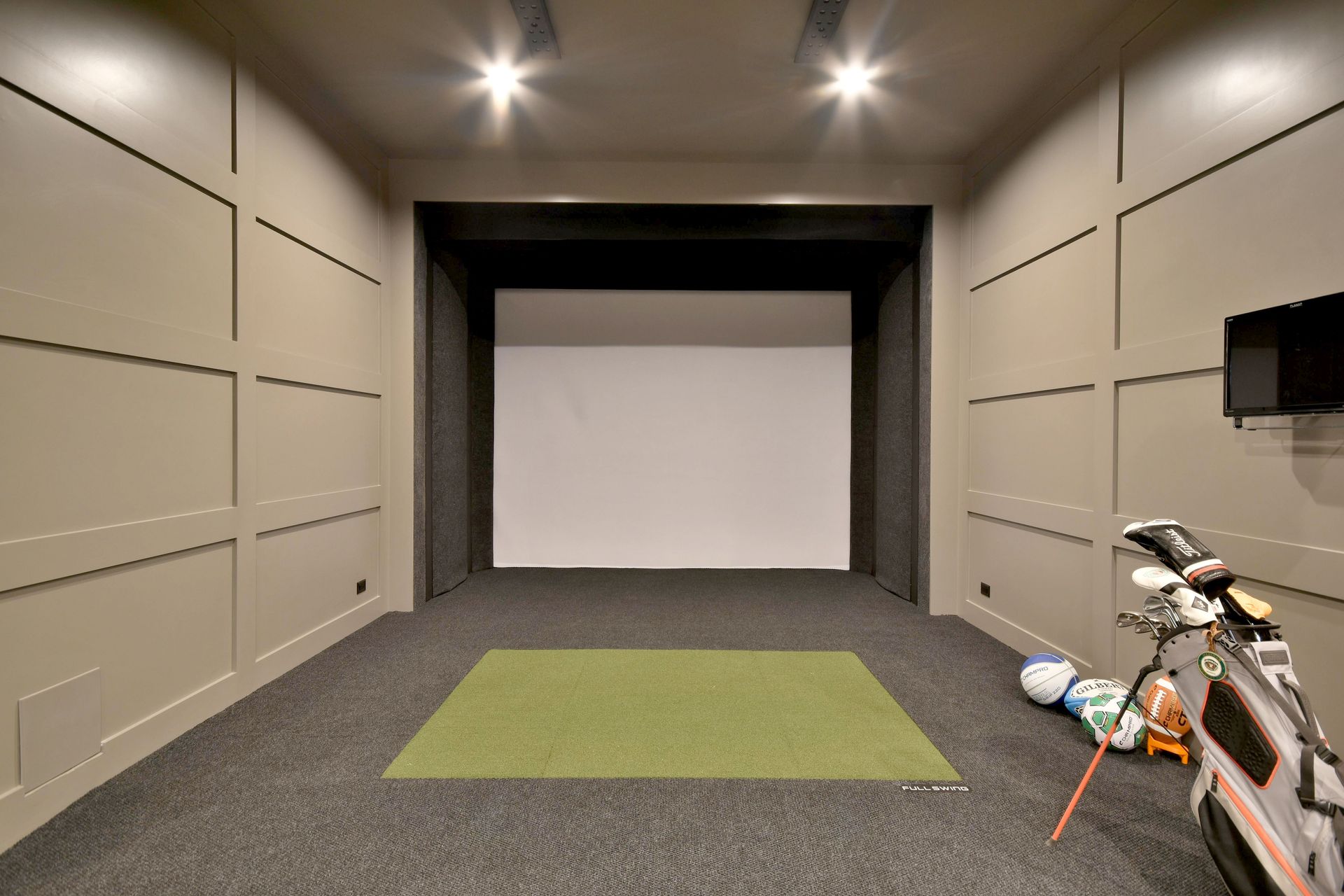 Indoor golf simulator room with a green hitting mat and golf clubs on the right.