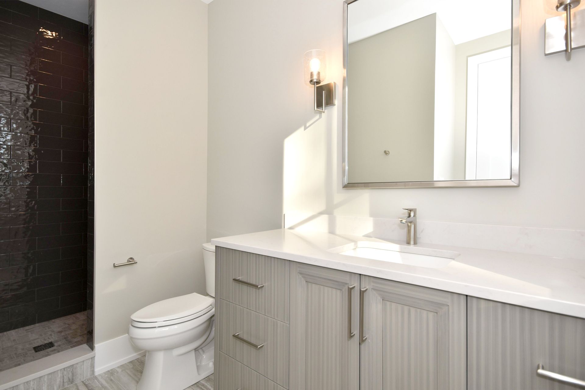 Modern bathroom with gray vanity, white countertop, mirror, sink, and toilet beside a dark shower.