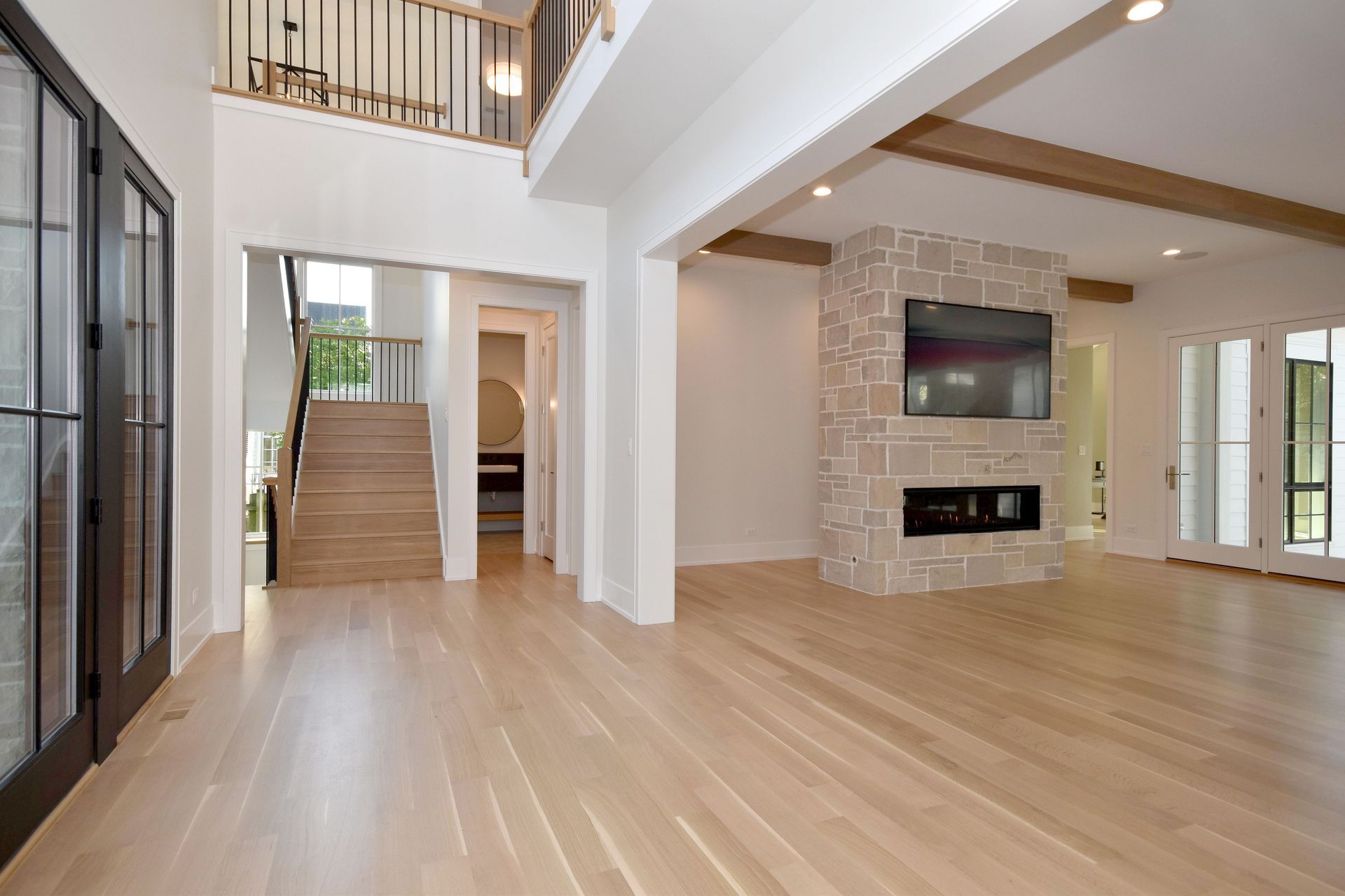 Spacious modern living room with light wood floors, stone fireplace, staircase, and large windows