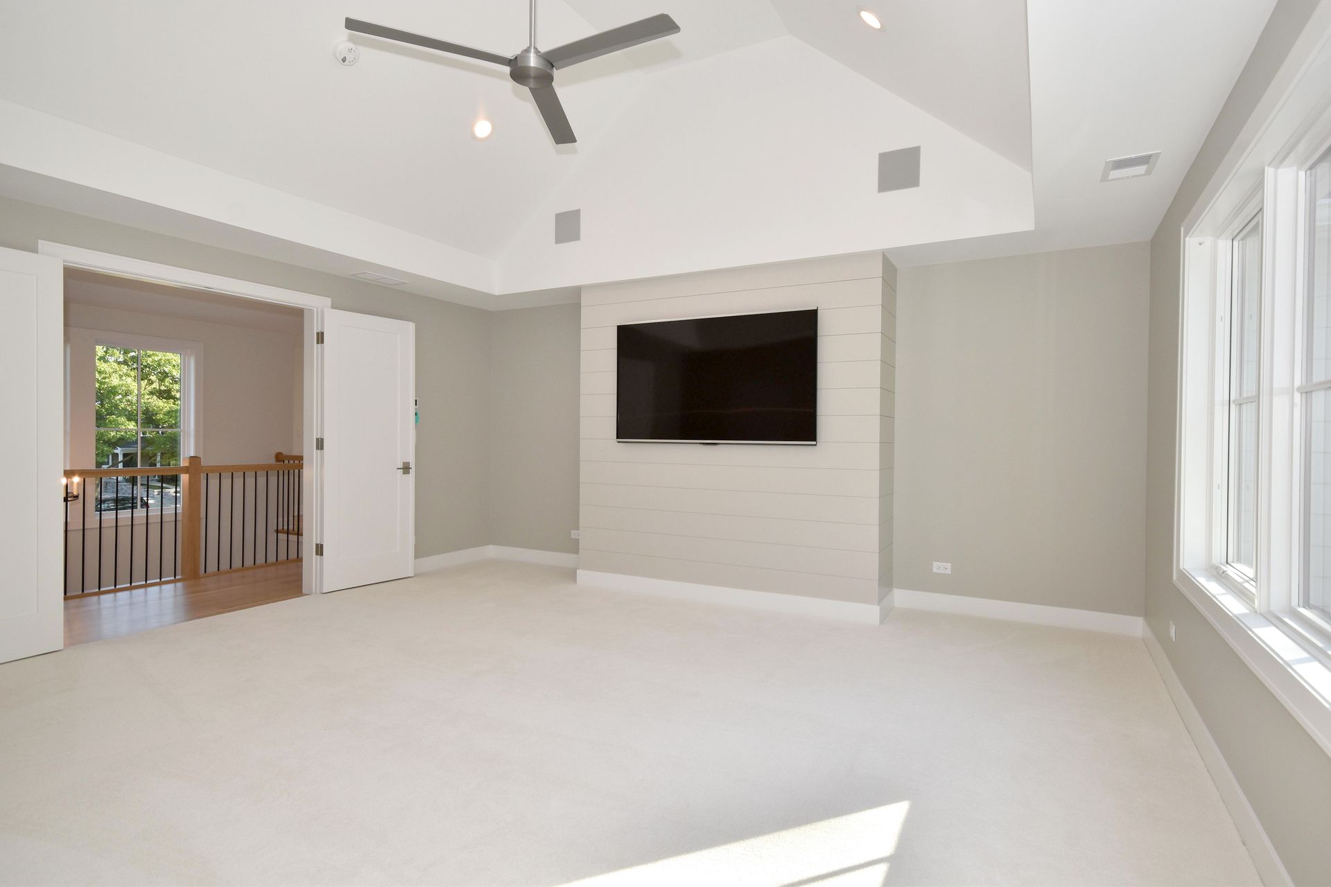 Bright living room with wall-mounted TV, ceiling fan, white carpet, and large windows.