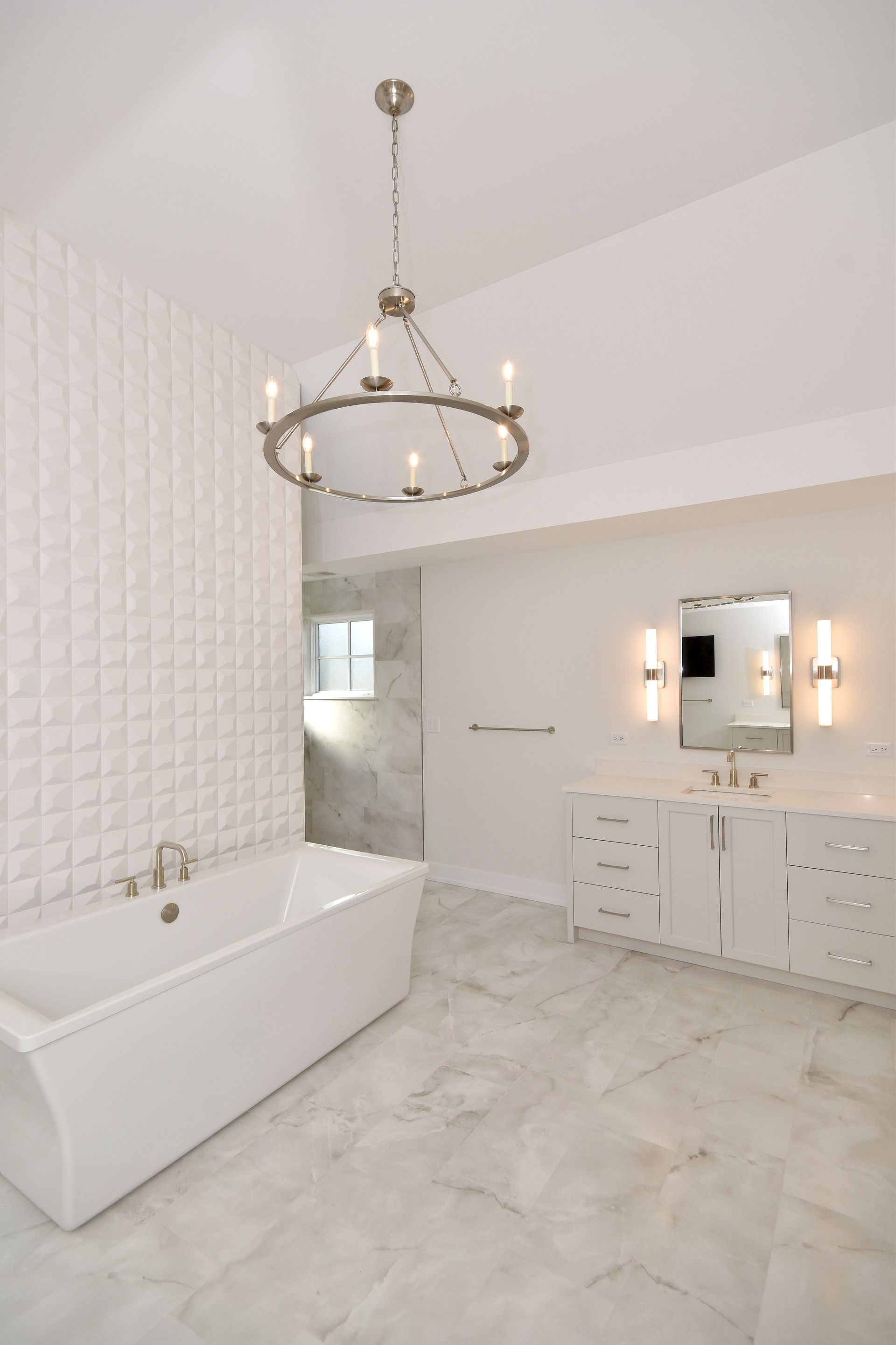 Modern white bathroom with freestanding tub, gold chandelier, patterned tile wall, and vanity mirror.