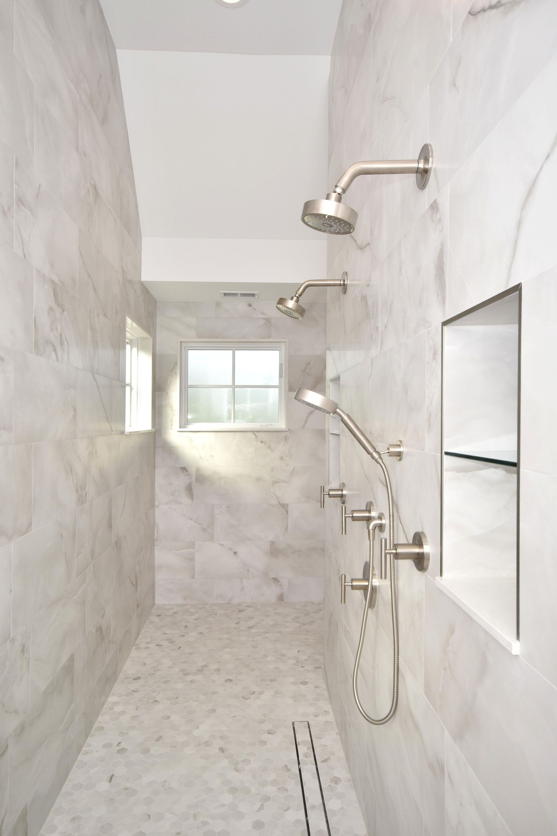 Tiled walk-in shower with grab bars, multiple showerheads, and a built-in shelf beside a frosted window.