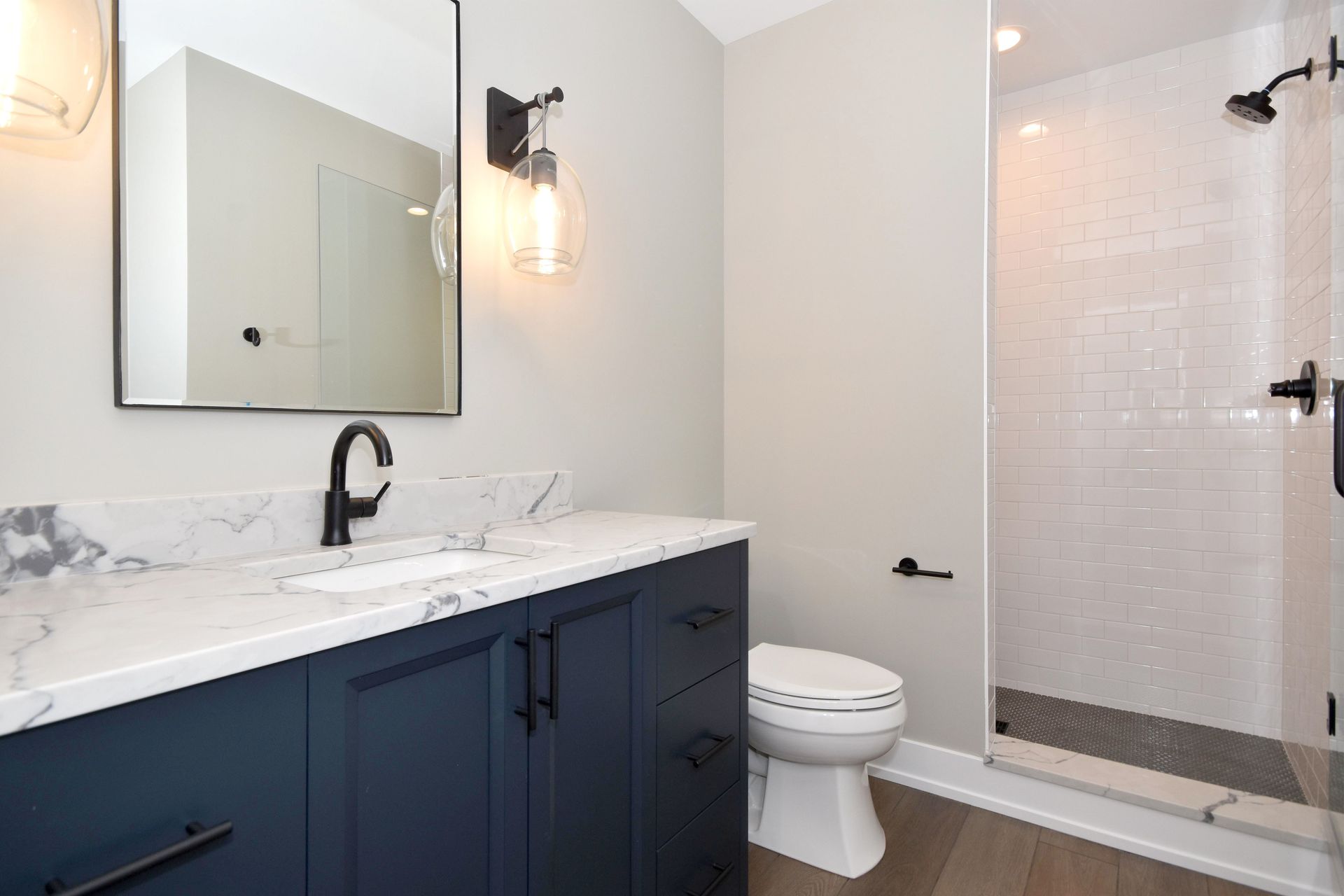 Modern bathroom with navy vanity, marble countertop, mirror, toilet, wall sconce, and glass shower