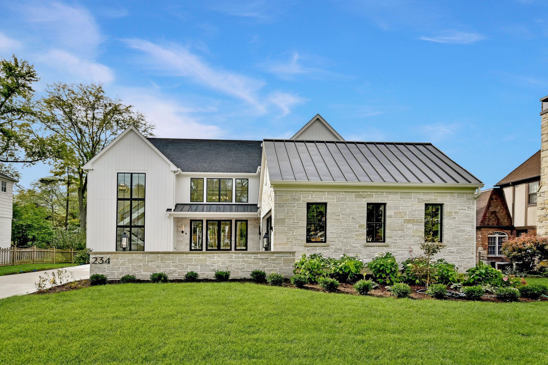 Modern two-story stone house with a metal roof and manicured front lawn under a blue sky