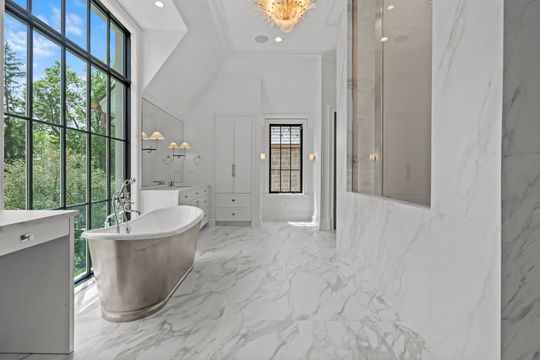 Elegant white marble bathroom with freestanding tub, large window, and gold chandelier
