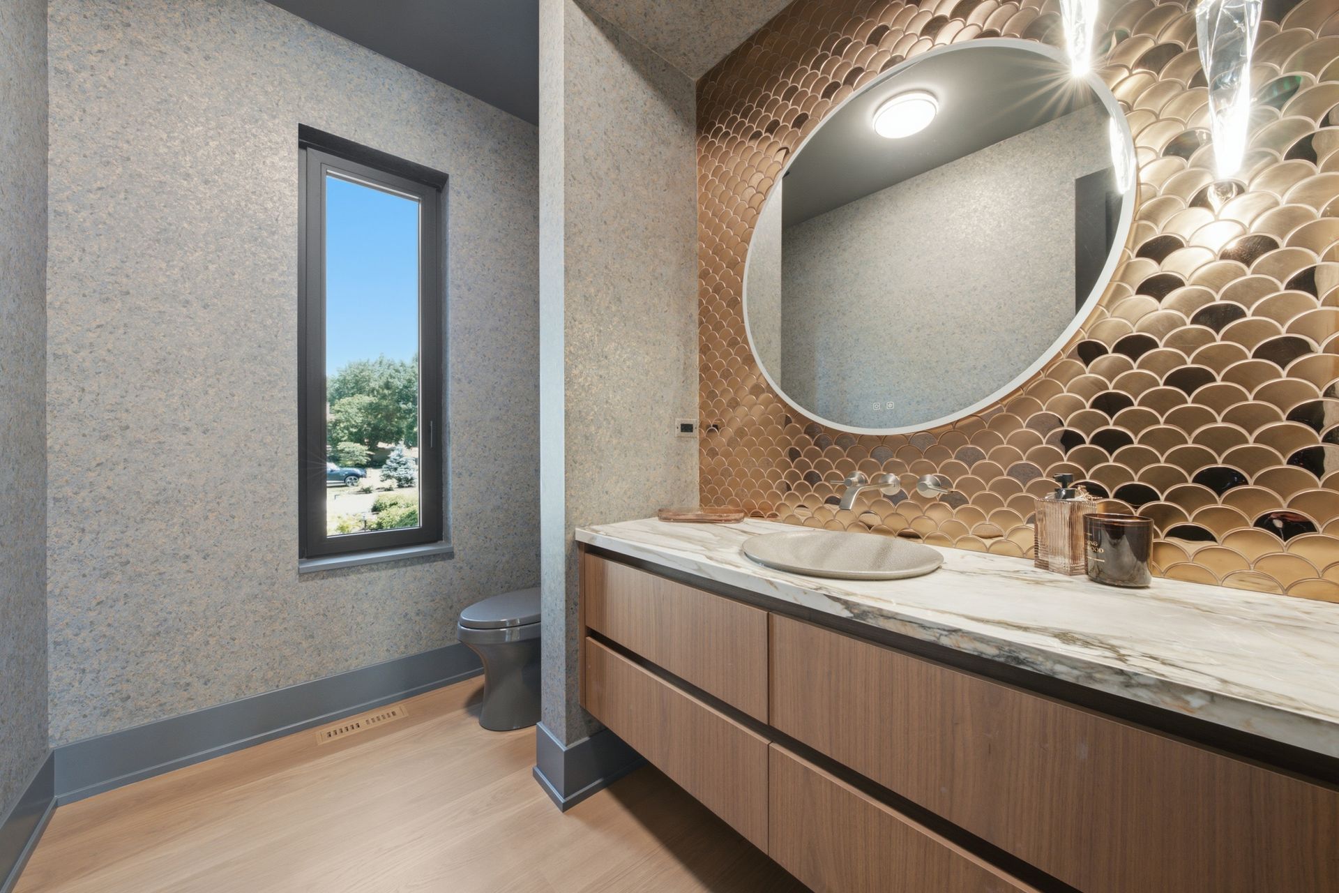 Modern bathroom vanity with round mirror, stone countertop, and window beside textured walls