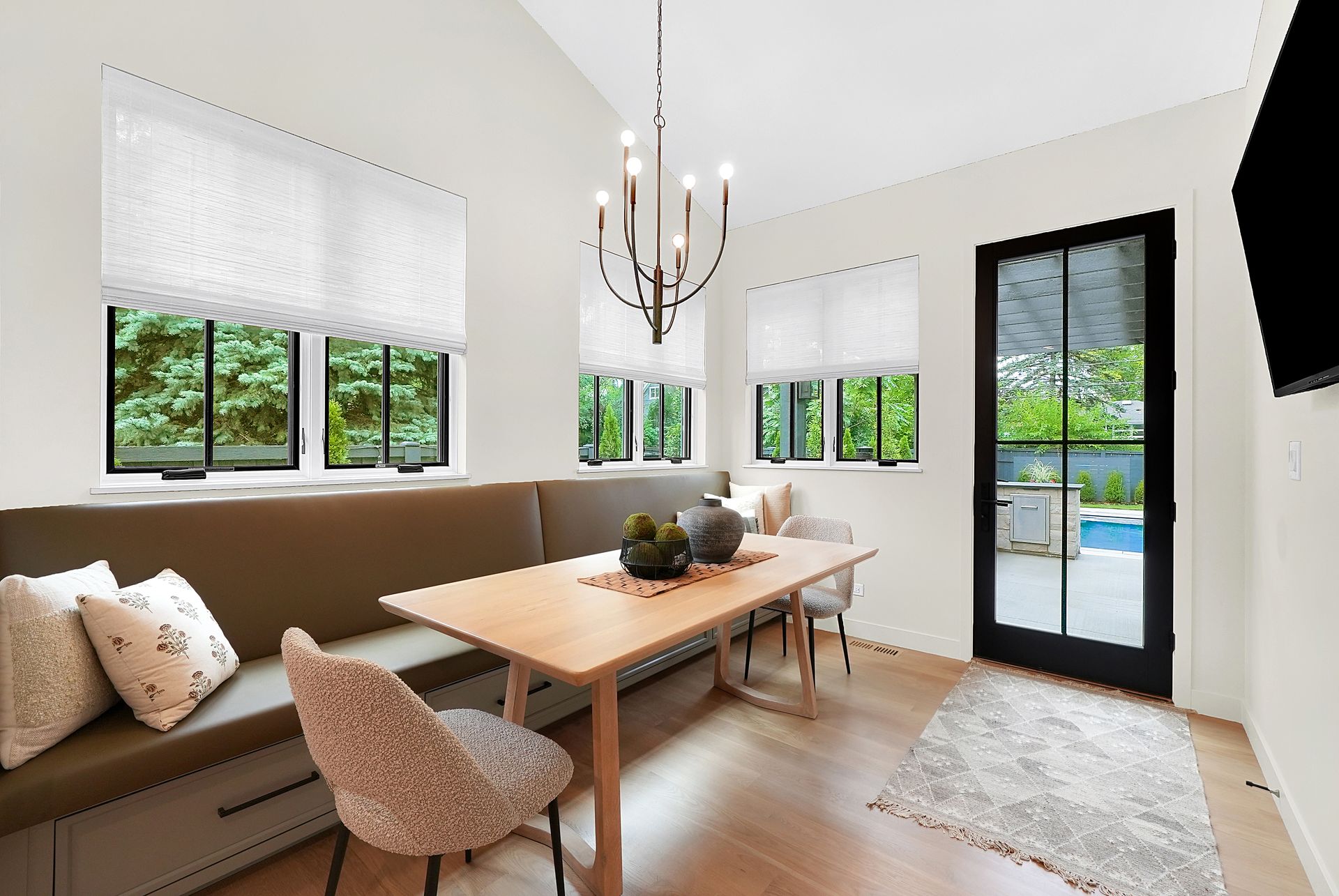 Bright dining room with large windows, wooden table, beige bench, chandelier, and a view of a pool outside