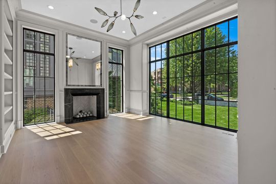 Bright empty living room with fireplace, white walls, and large black-framed windows overlooking a green yard