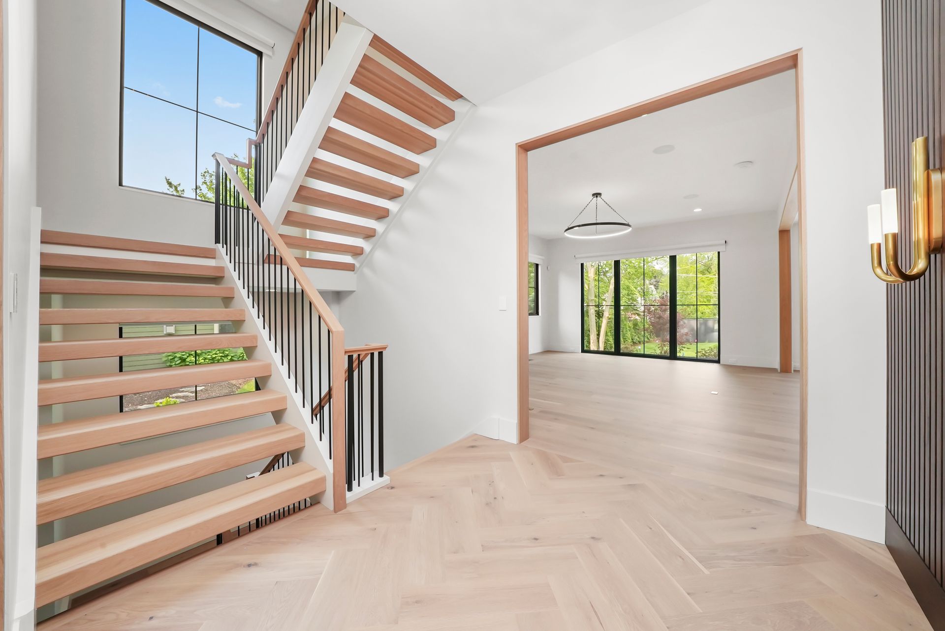 Modern entry hall with floating wooden stairs, white walls, and herringbone wood floor leading to a bright room