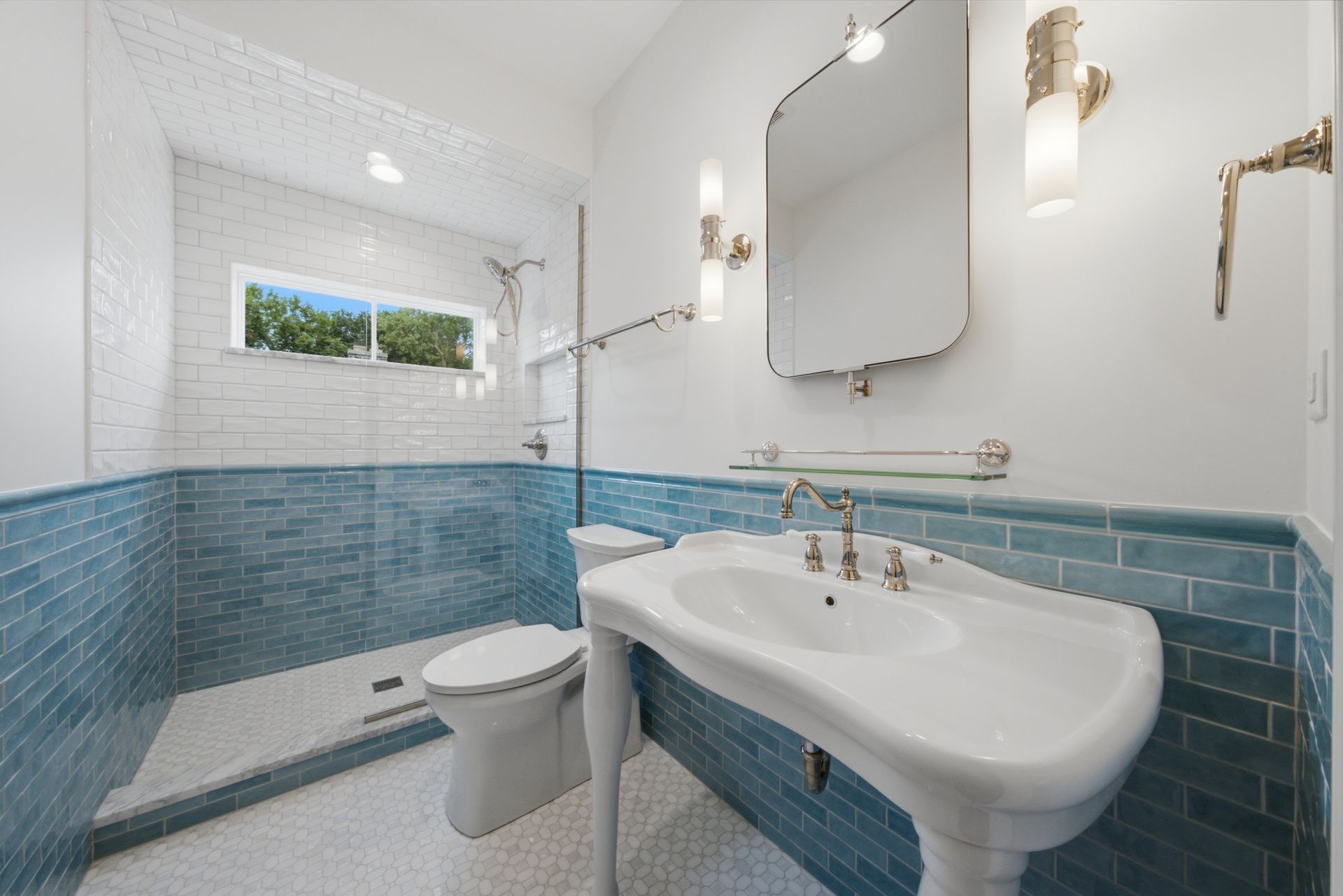 Bright white bathroom with blue tile, a pedestal sink, toilet, mirror, and a small window.