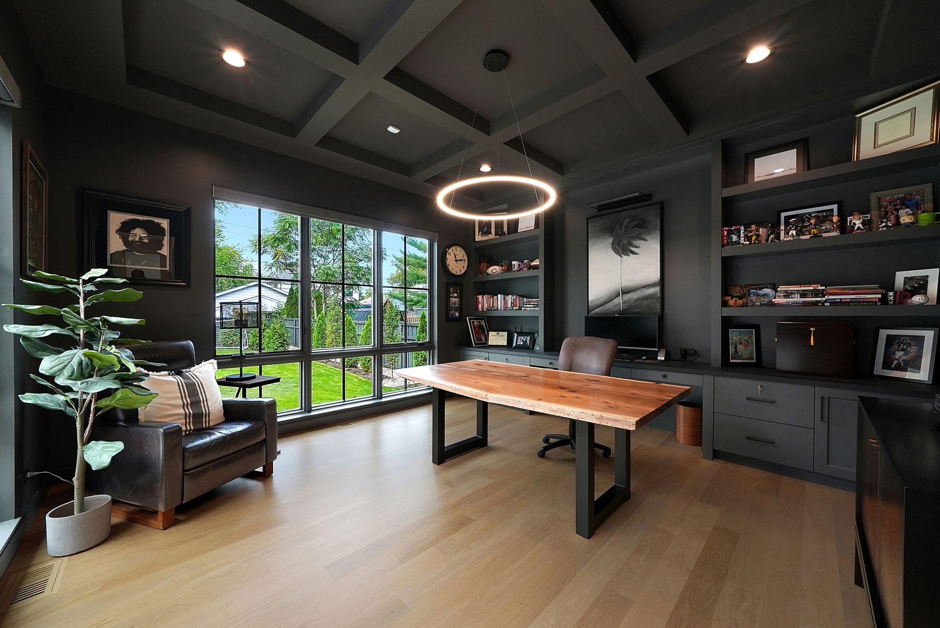 Modern home office with a wood desk, dark walls, large windows, and indoor plants