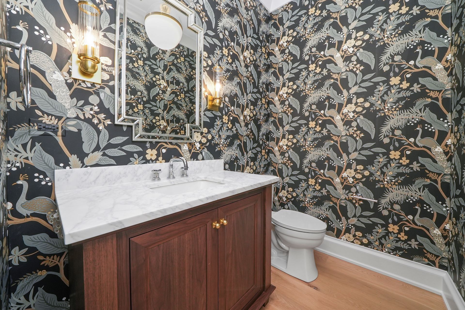 Small bathroom with floral wallpaper, white vanity, gold mirror, and toilet on wood floor