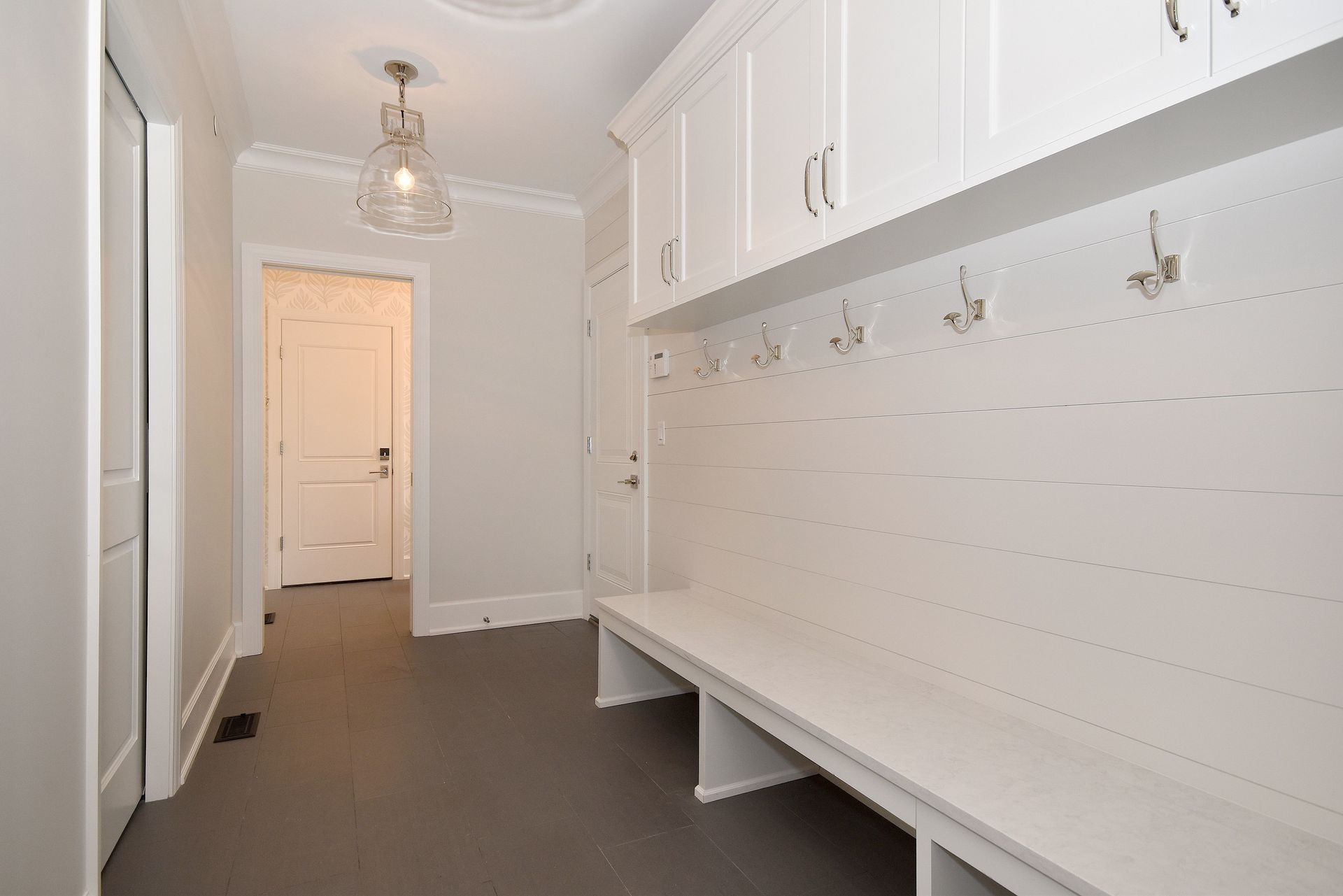 Narrow white mudroom with built-in bench, hooks, cabinets, and a door at the end.