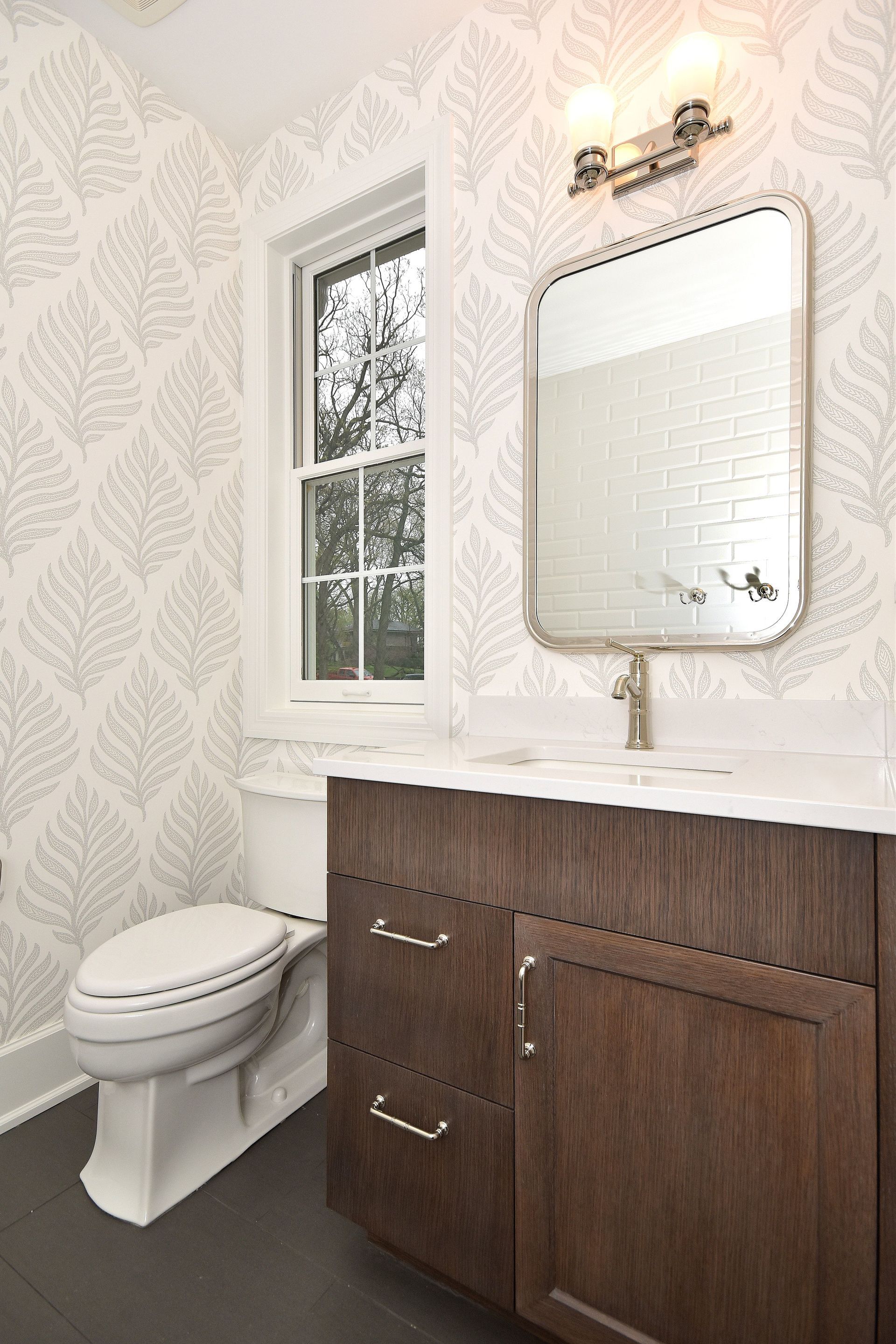 Bright bathroom with white patterned wallpaper, window, wall mirror, and dark wood vanity next to a toilet.