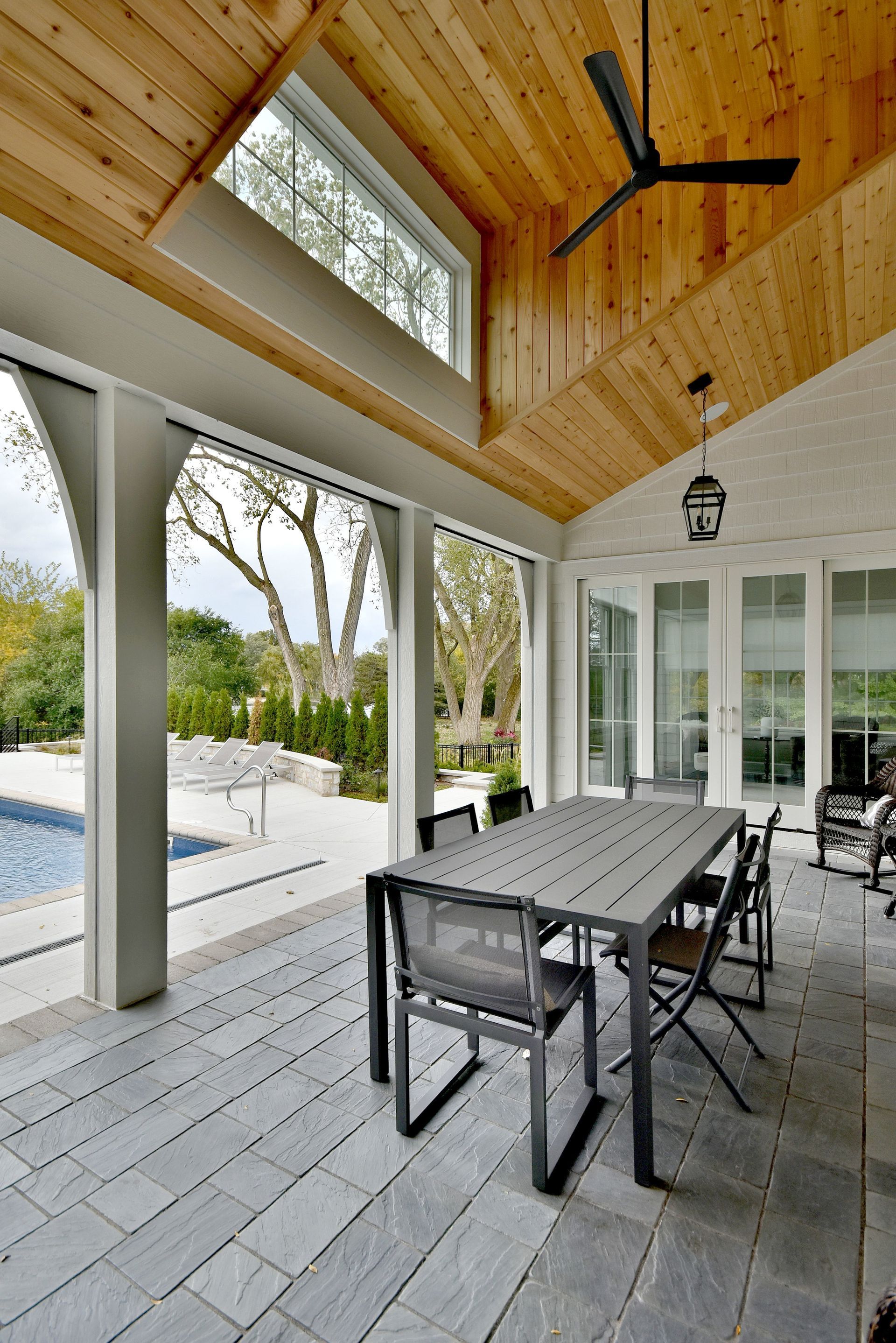 Covered patio with a long dining table, chairs, and ceiling fan beside a pool and glass doors