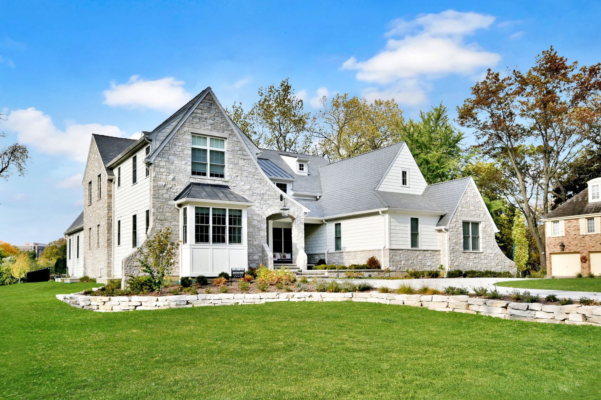 Large modern stone house with white trim, gabled roofs, and a manicured green lawn under a blue sky