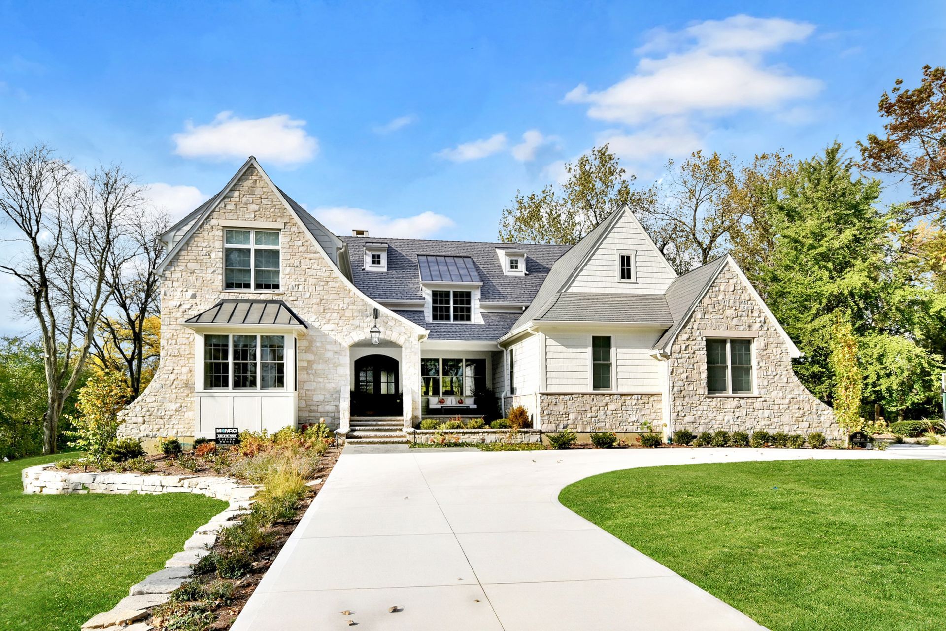 Stone suburban house with white trim, front lawn, and a curved concrete driveway under a blue sky