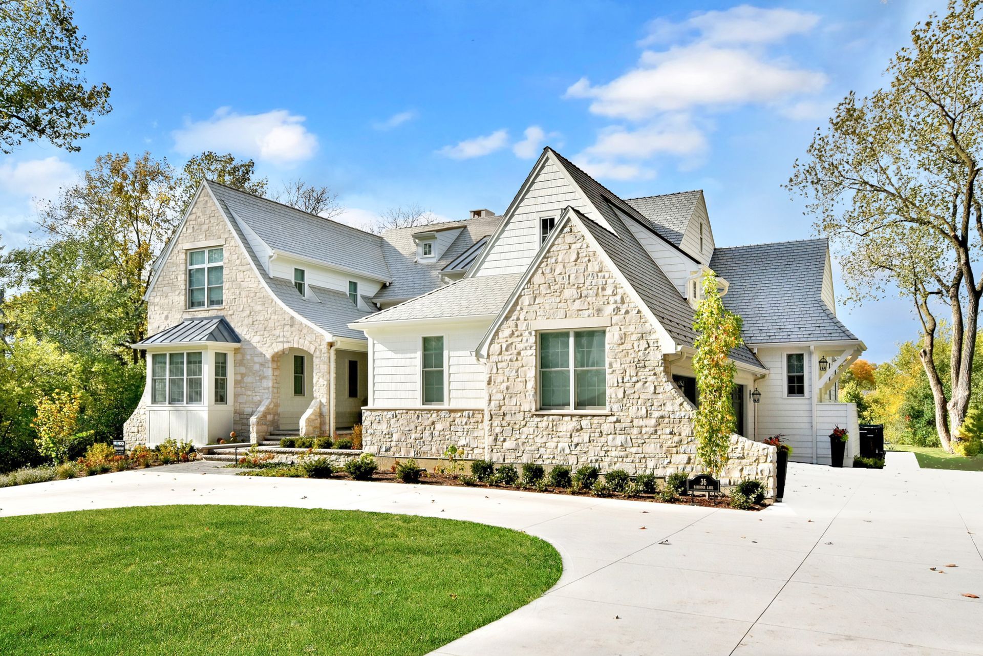 Stone-faced suburban house with gabled roof, green lawn, and curved driveway under a blue sky
