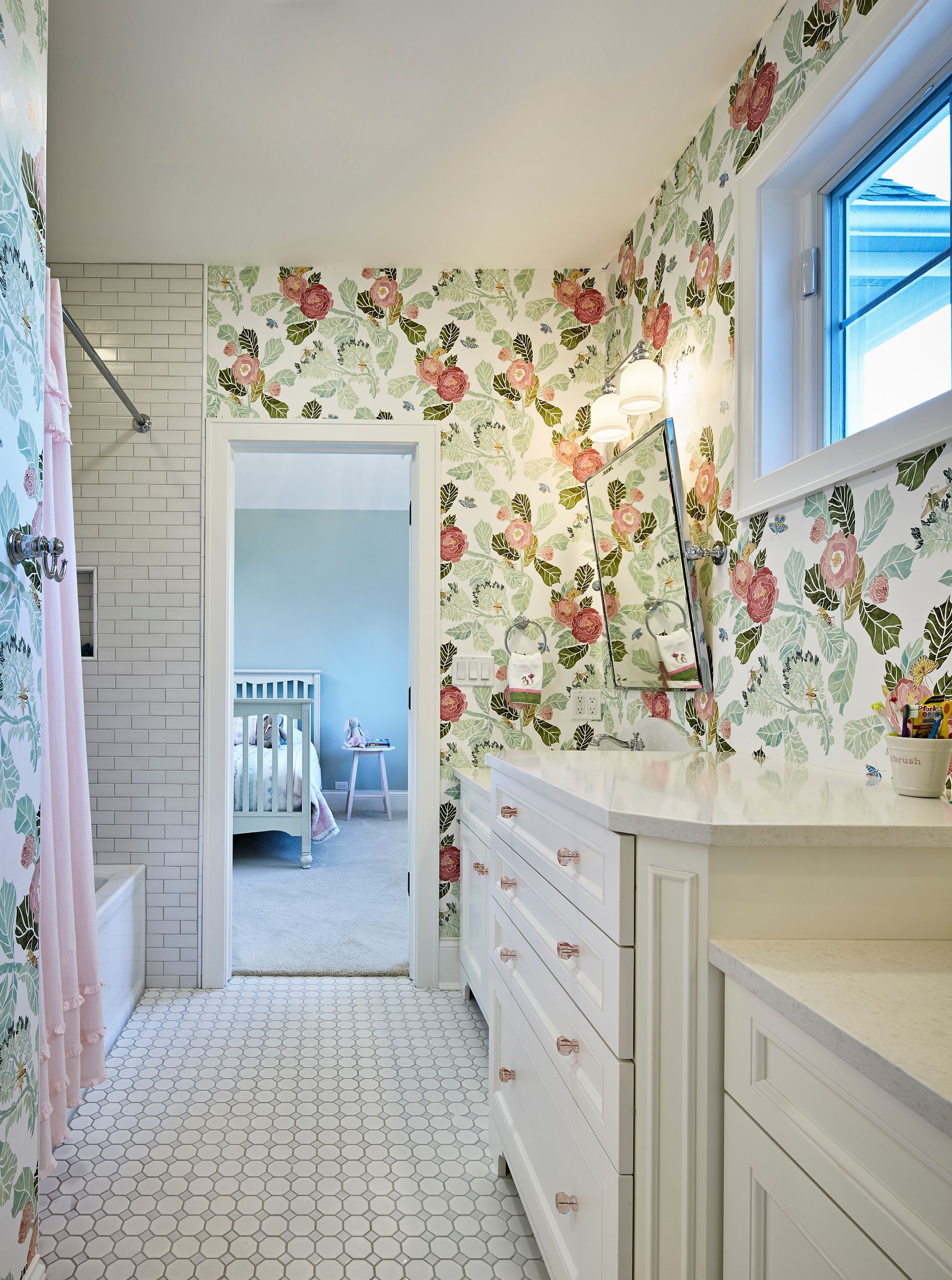 Bright floral wallpaper in a narrow bathroom with white cabinets, a shower, and a window.