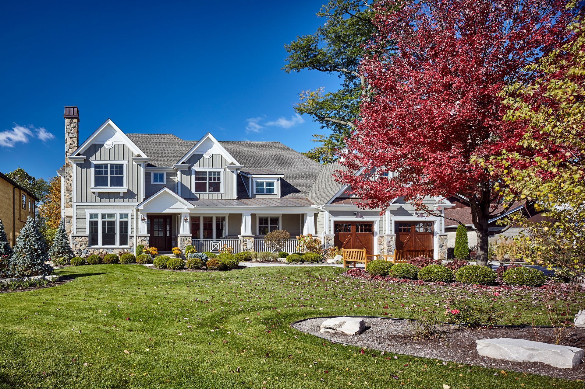 Suburban house with white siding, garage, and a large red tree beside a green lawn under a blue sky