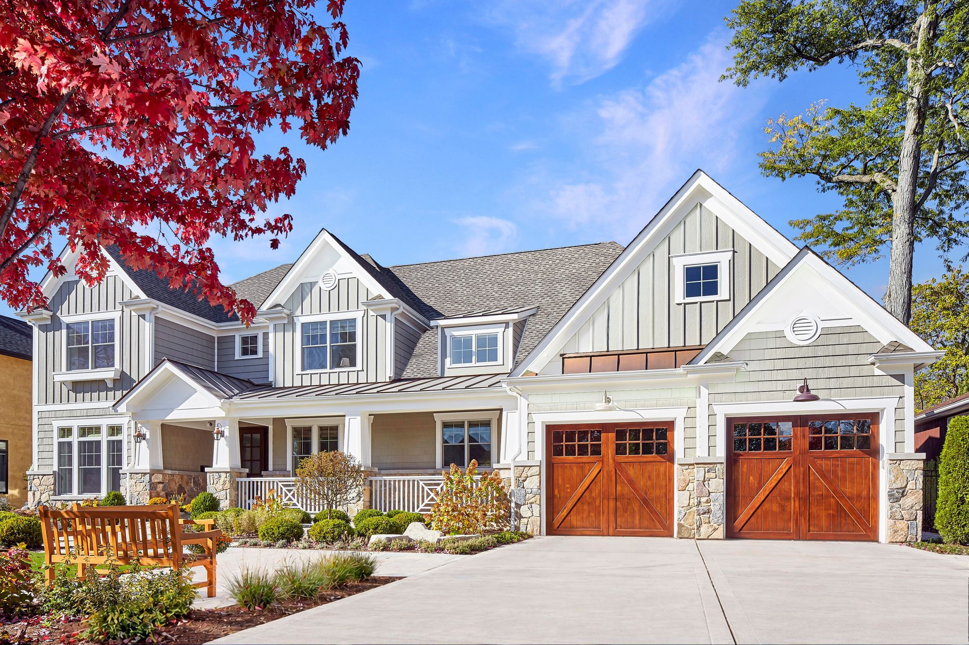 Large white suburban house with two wooden garage doors, front porch, and red and green trees.