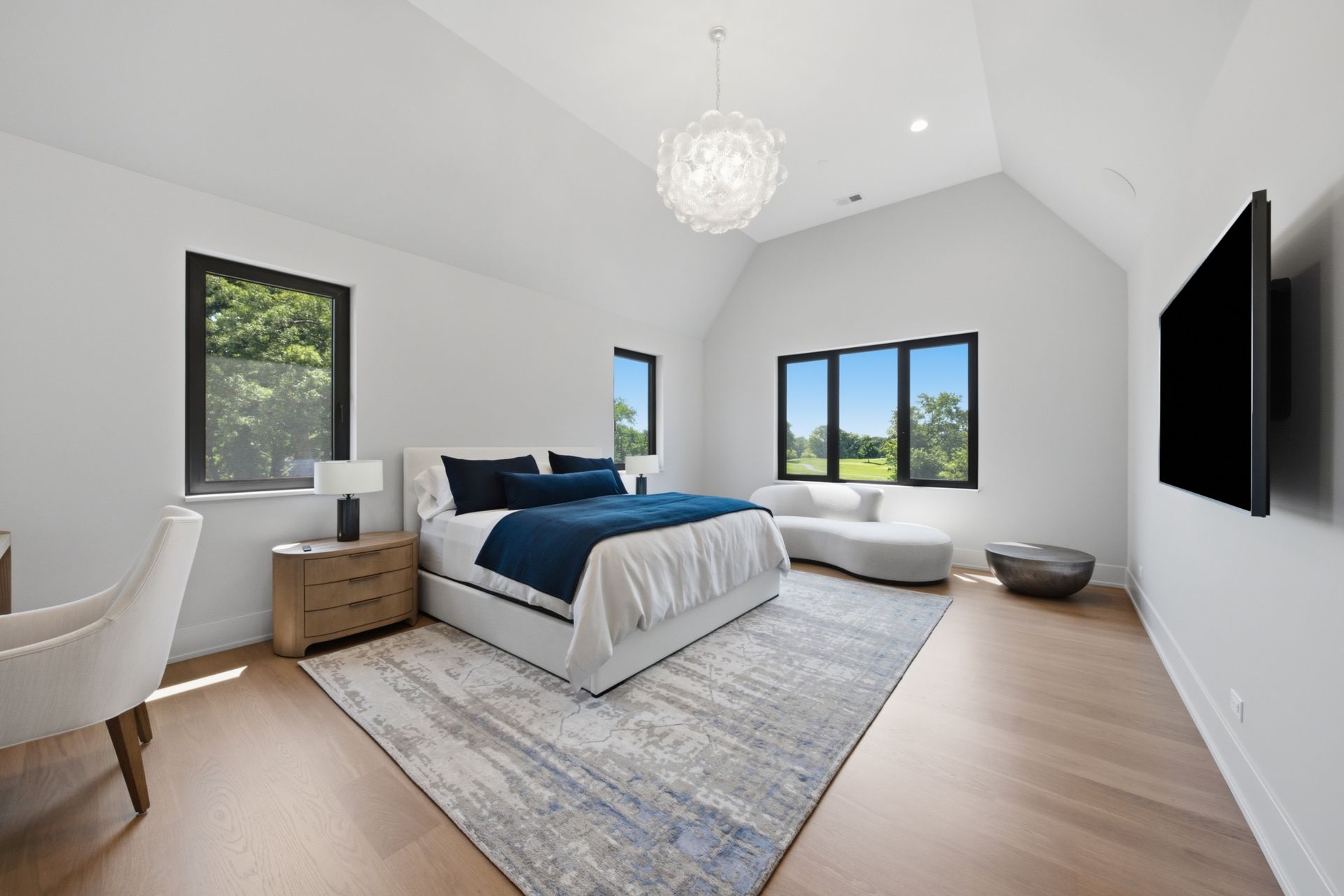 Bright modern bedroom with white walls, blue bedding, large windows, and a wall-mounted TV.