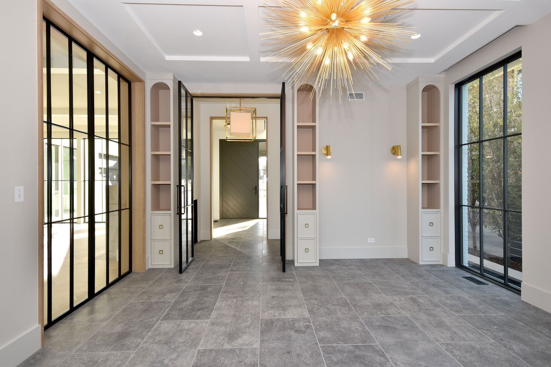 Elegant empty hallway with gray carpet, gold starburst chandelier, and glass doors leading to a bright room