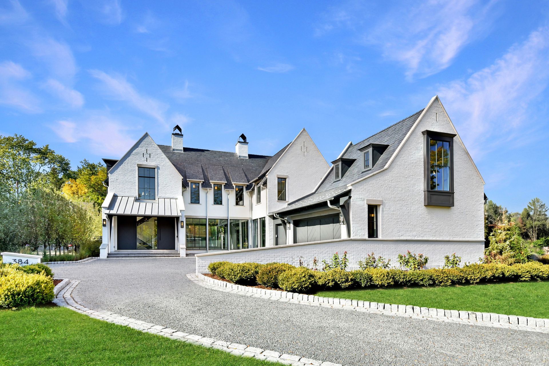 Large gray stone house with a curved driveway, manicured lawn, and blue sky