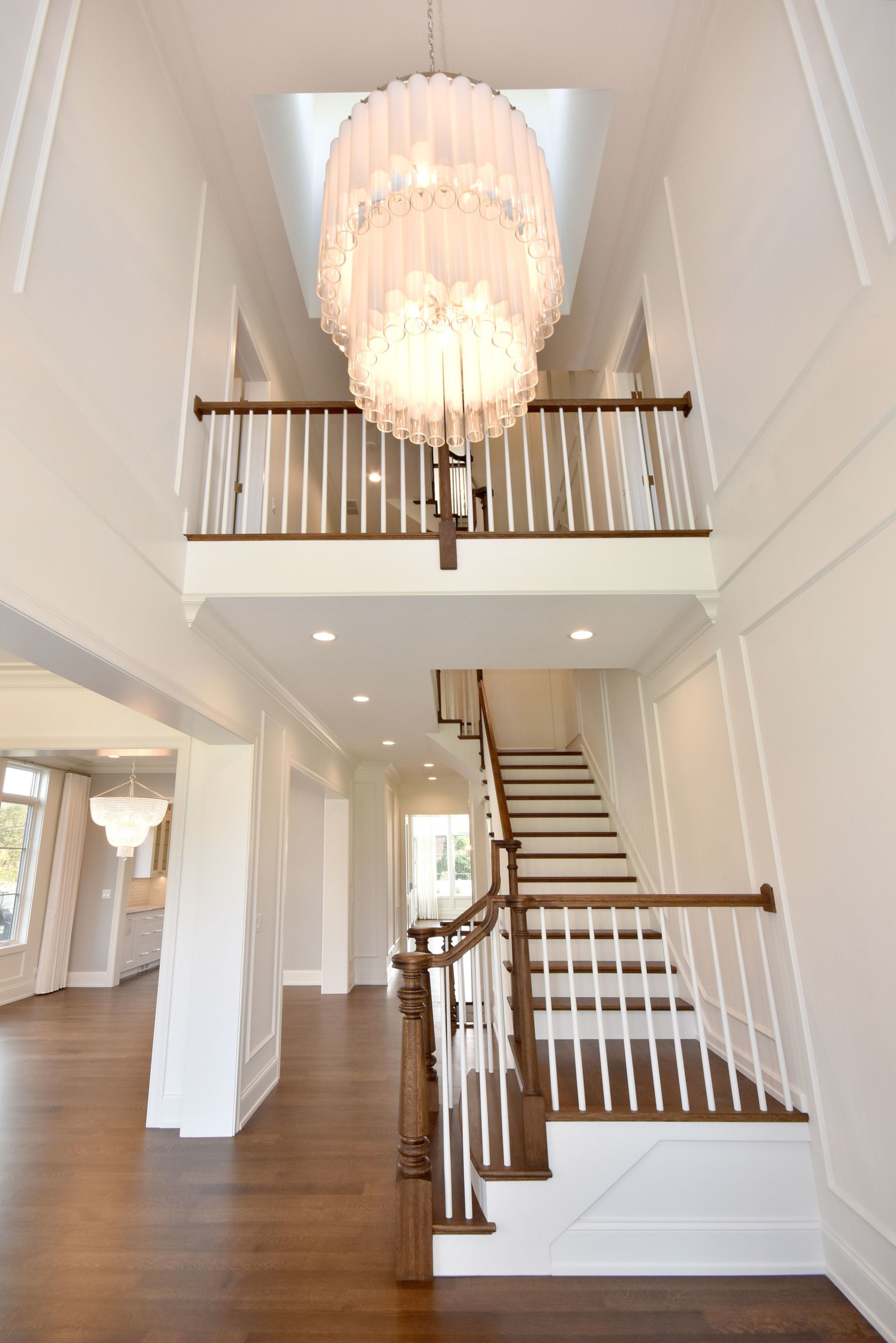 Bright foyer with white staircase, wood floors, and a large chandelier under a loft balcony