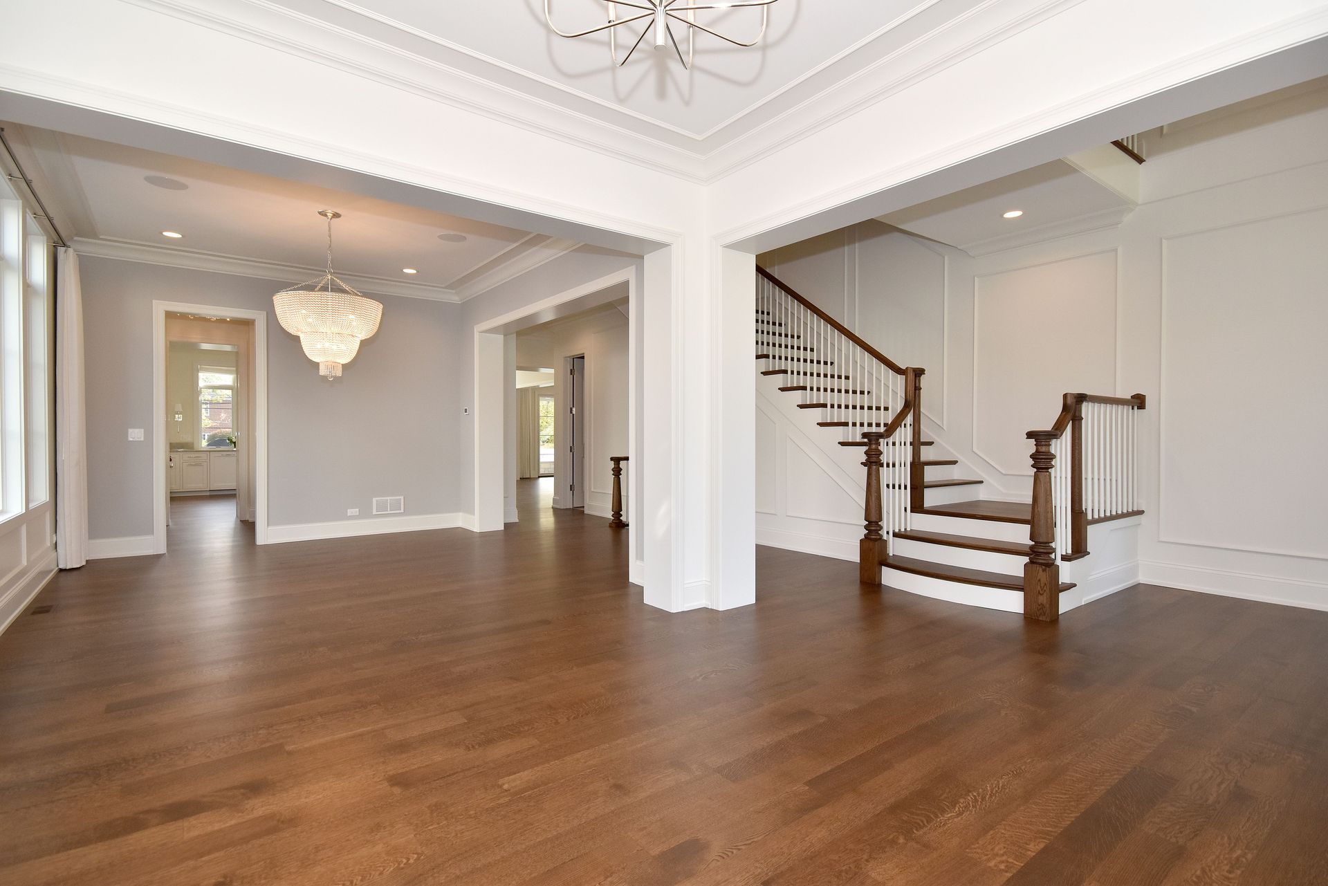 Spacious empty living room with hardwood floors, white walls, chandeliers, and a staircase on the right.