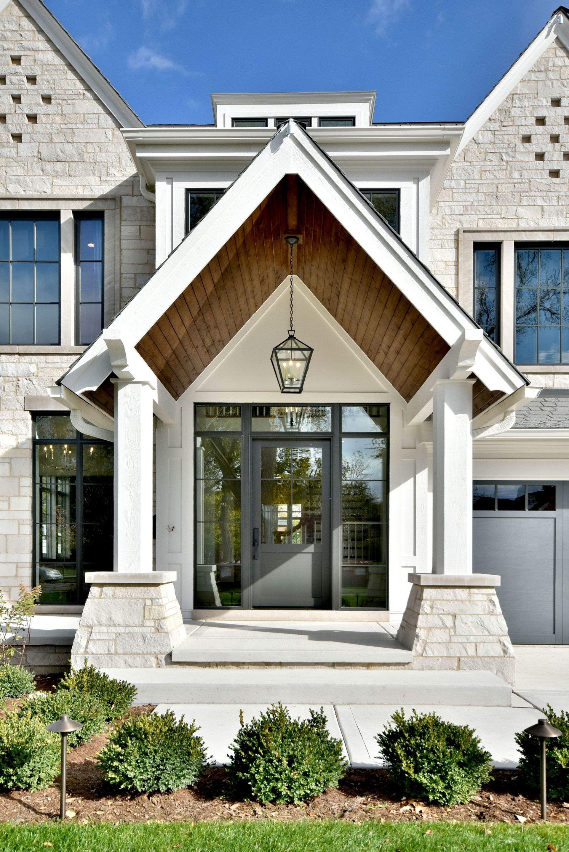 Modern house entrance with stone facade, peaked wood awning, glass double doors, and landscaped front shrubs
