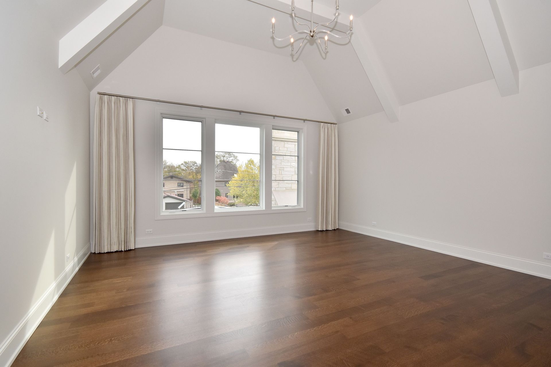 Empty bright attic room with hardwood floors, white walls, and a window with curtains