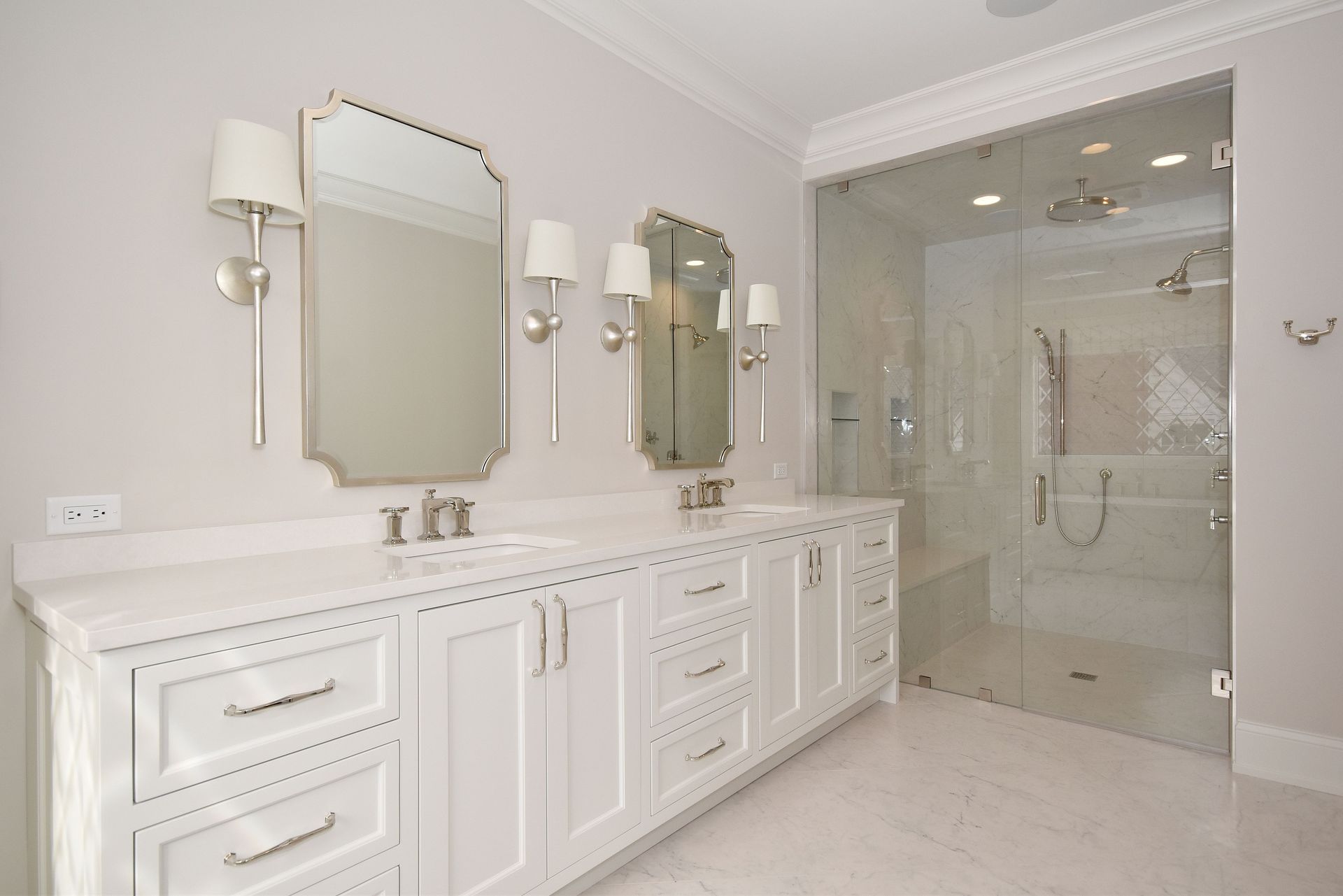 Elegant white bathroom with double vanity, mirrors, wall sconces, and glass shower enclosure