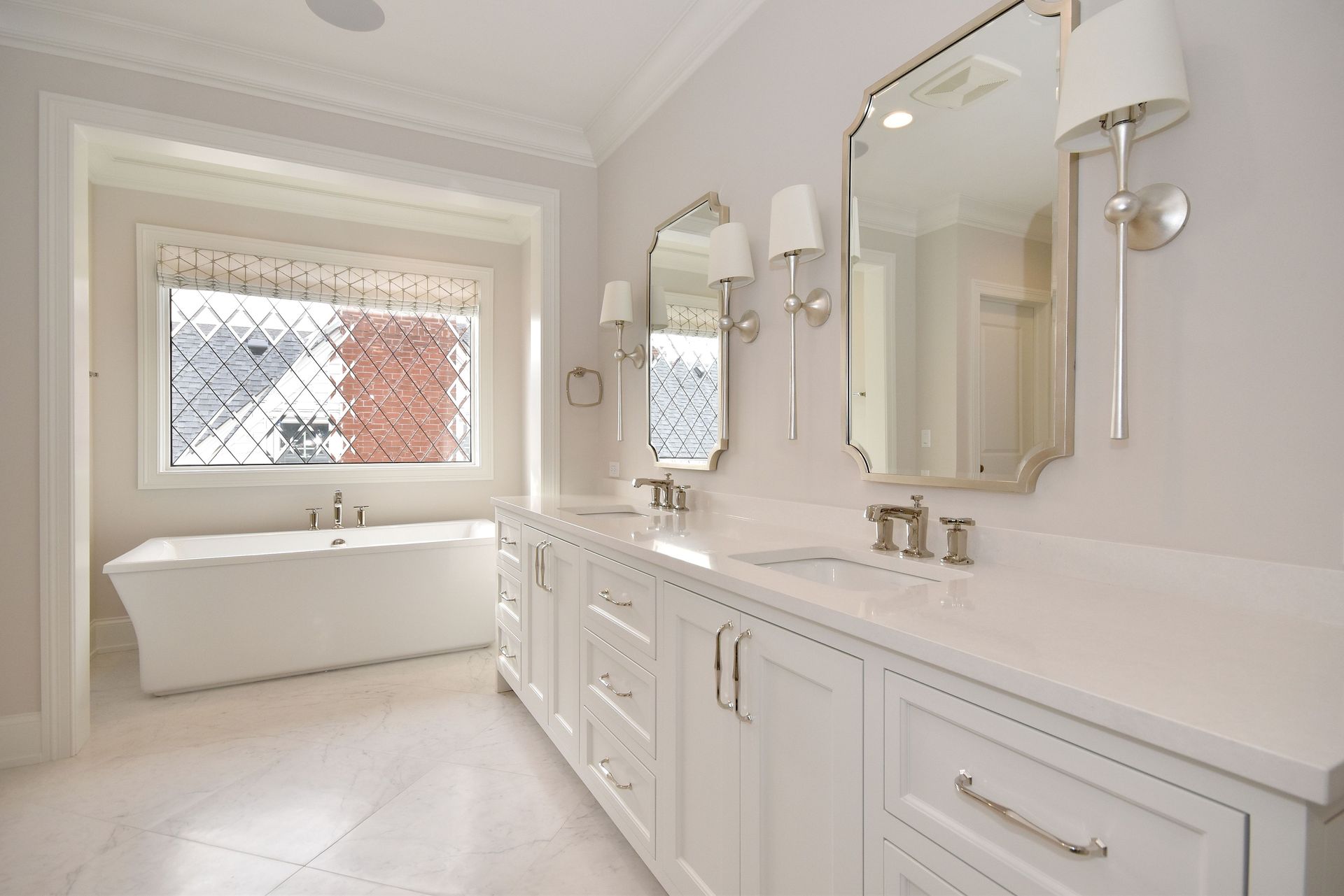 Bright white bathroom with double vanity, large mirror, wall sconces, and a built-in tub by a window.