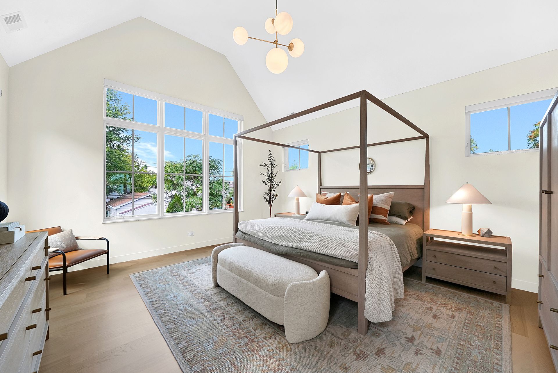Bright bedroom with four-poster bed, gray bench, large windows, and light wood furniture.