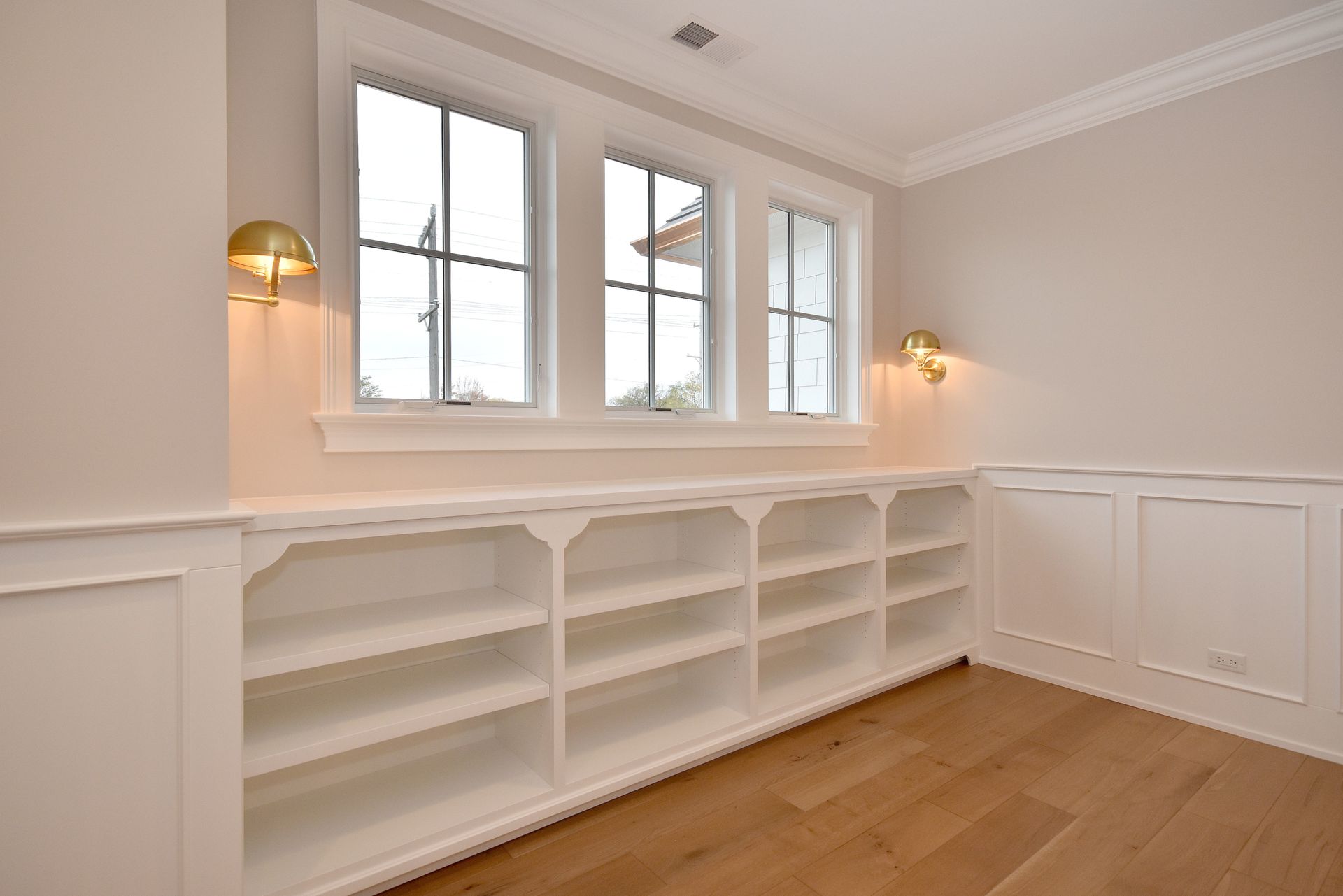 Bright empty room with white built-in shelves, windows, wall sconces, and hardwood floor