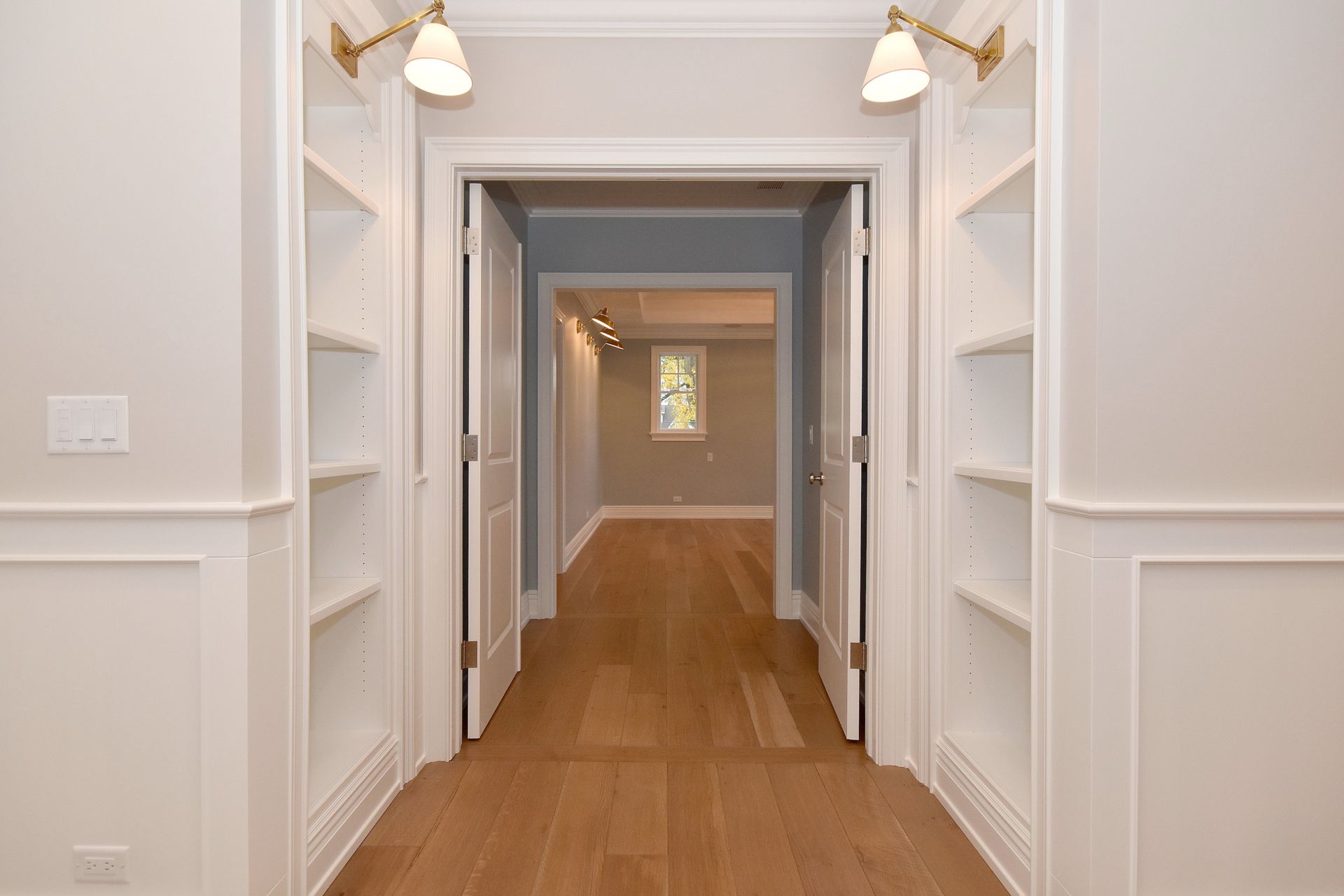 Bright hallway with white built-in shelves, wood floor, and lights leading to a doorway.
