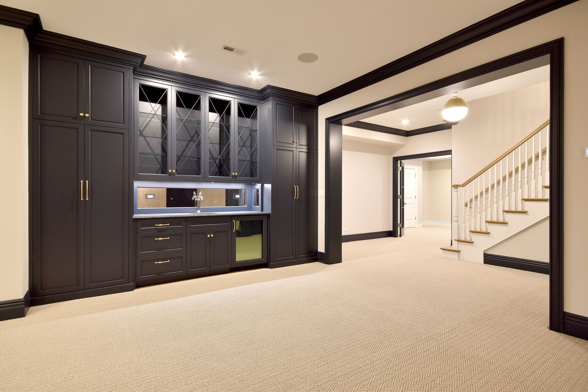 Modern basement with dark built-in cabinets, carpeted floor, and staircase leading to a bright hallway
