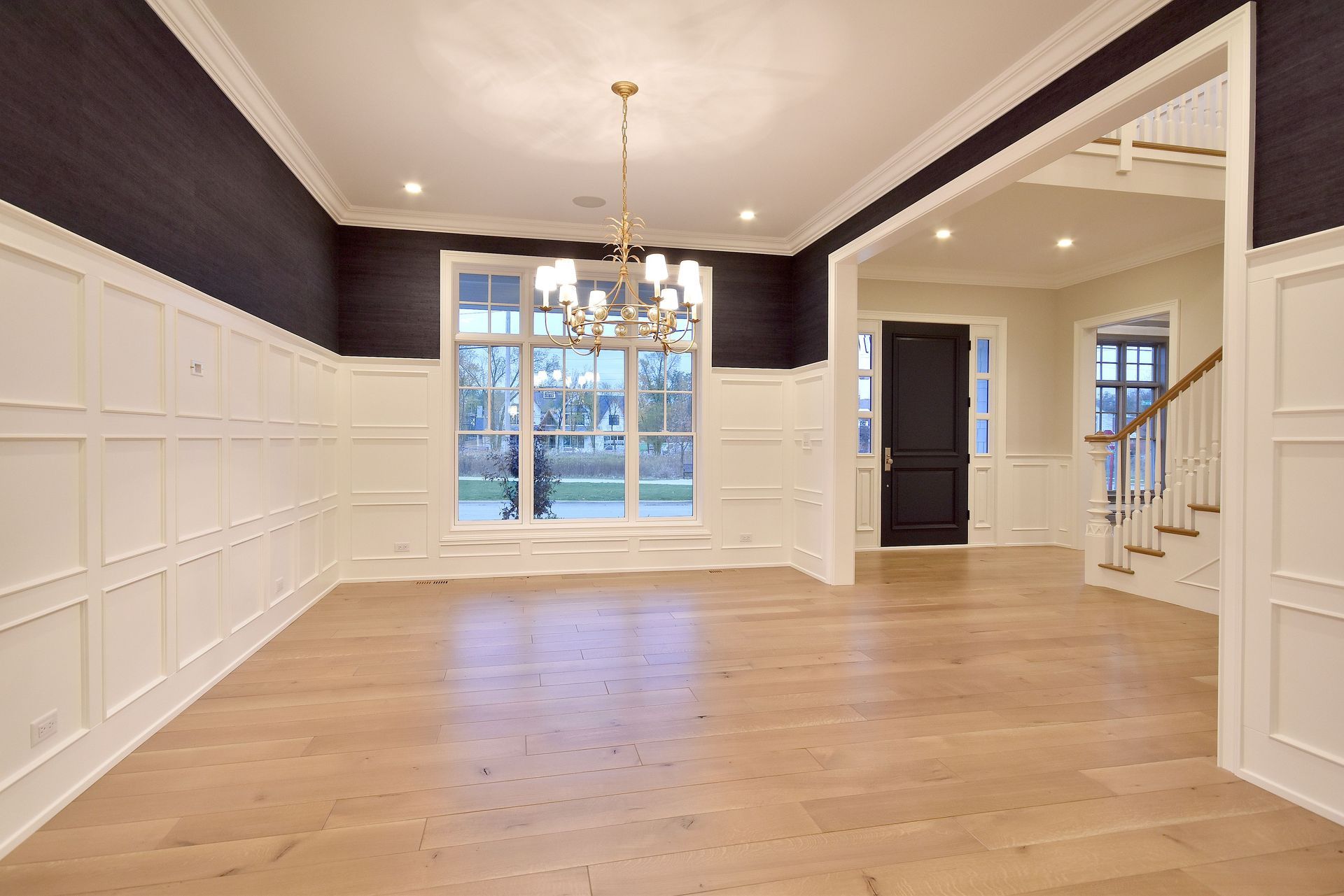 Empty dining room with light wood floors, white wainscoting, dark accent walls, and a chandelier.