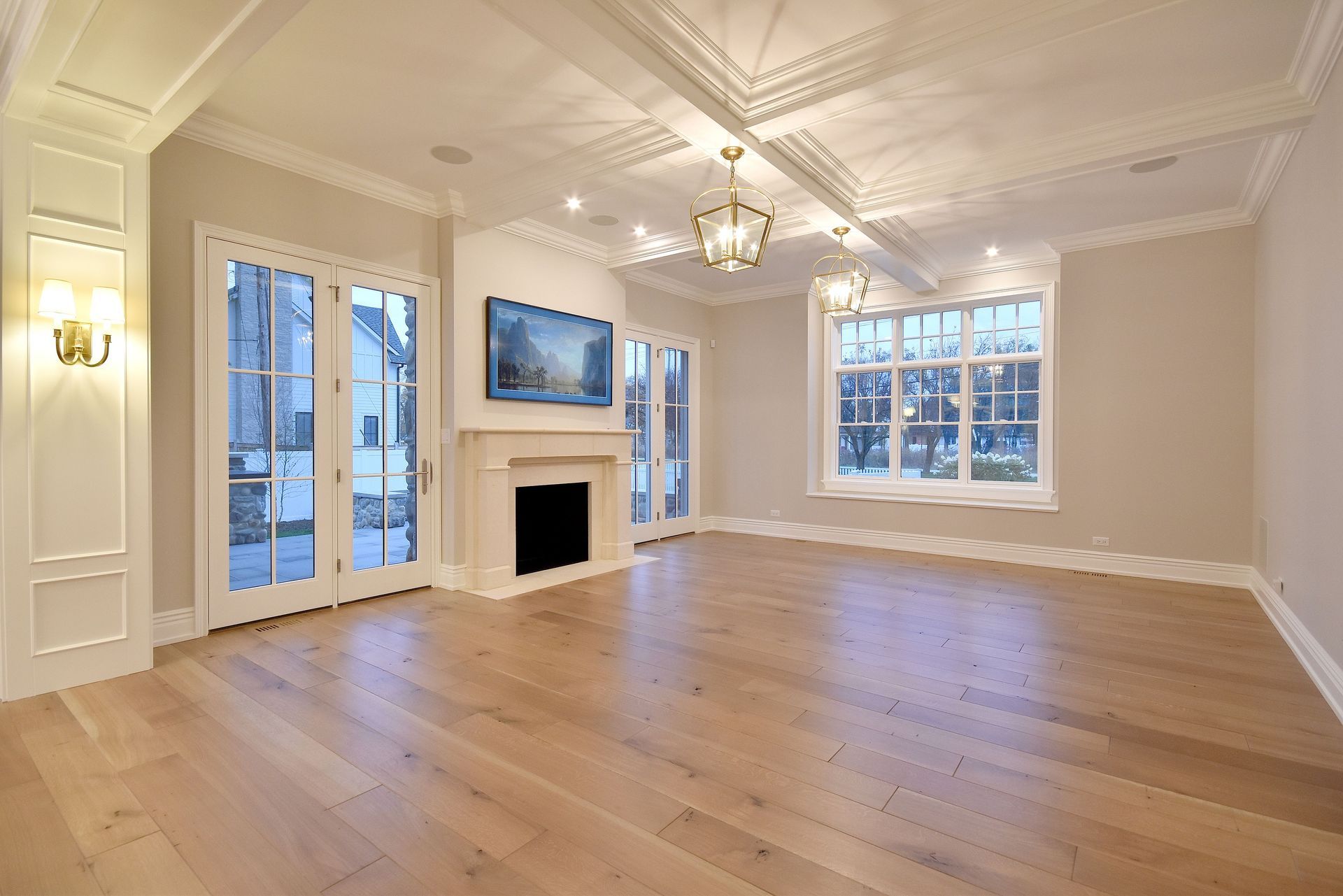 Bright empty living room with hardwood floors, fireplace, chandelier, wall-mounted TV, and large windows.