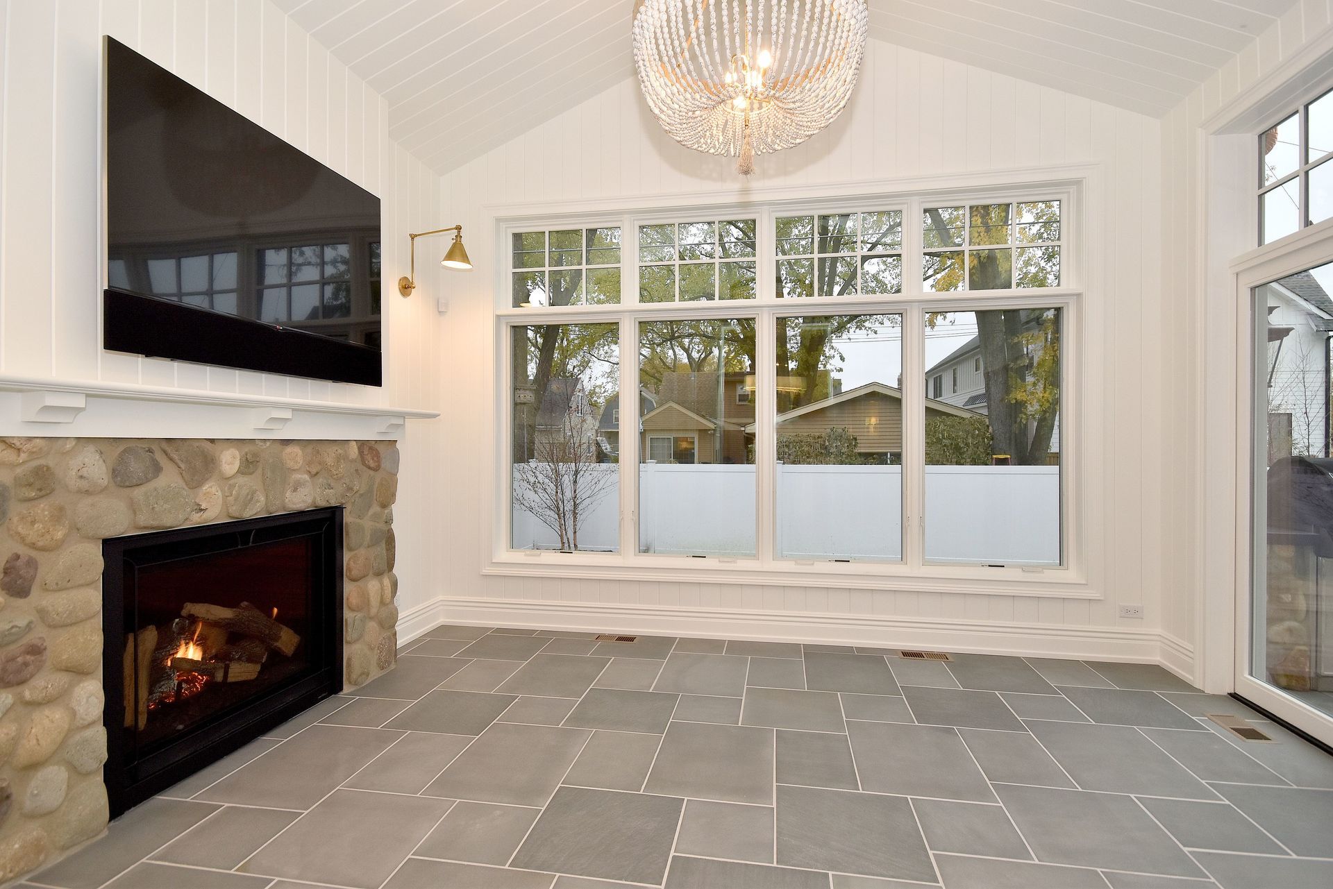 Bright living room with stone fireplace, large windows, gray tile floor, and chandelier
