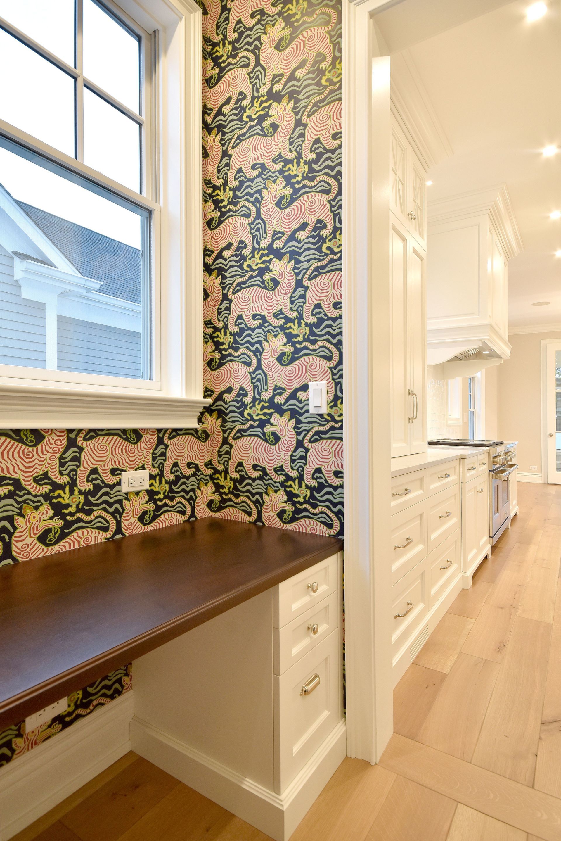 Built-in desk nook with floral wallpaper beside a bright white hallway kitchen.