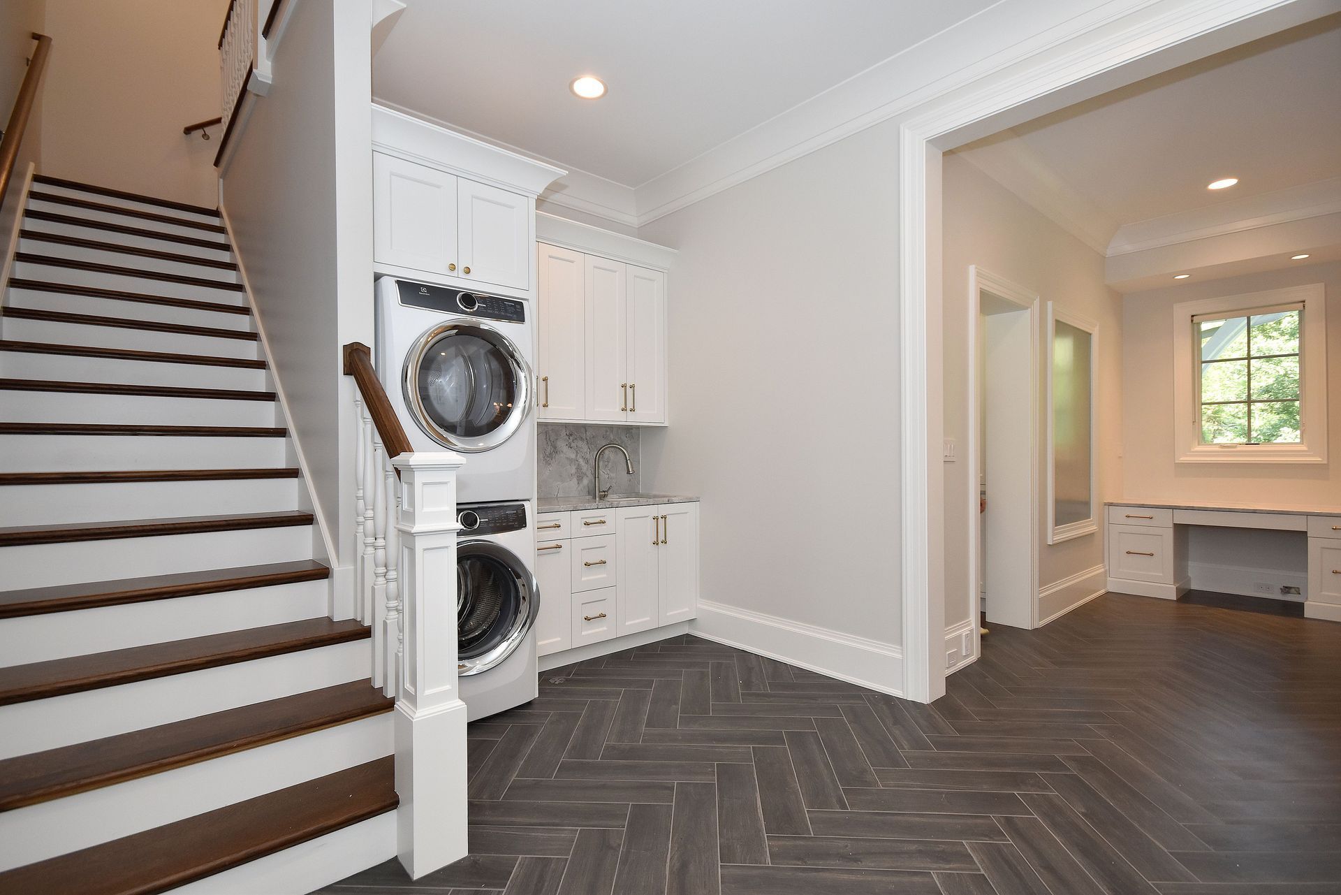 Bright foyer with staircase, stacked washer and dryer, dark herringbone floor, and adjacent room with window seat